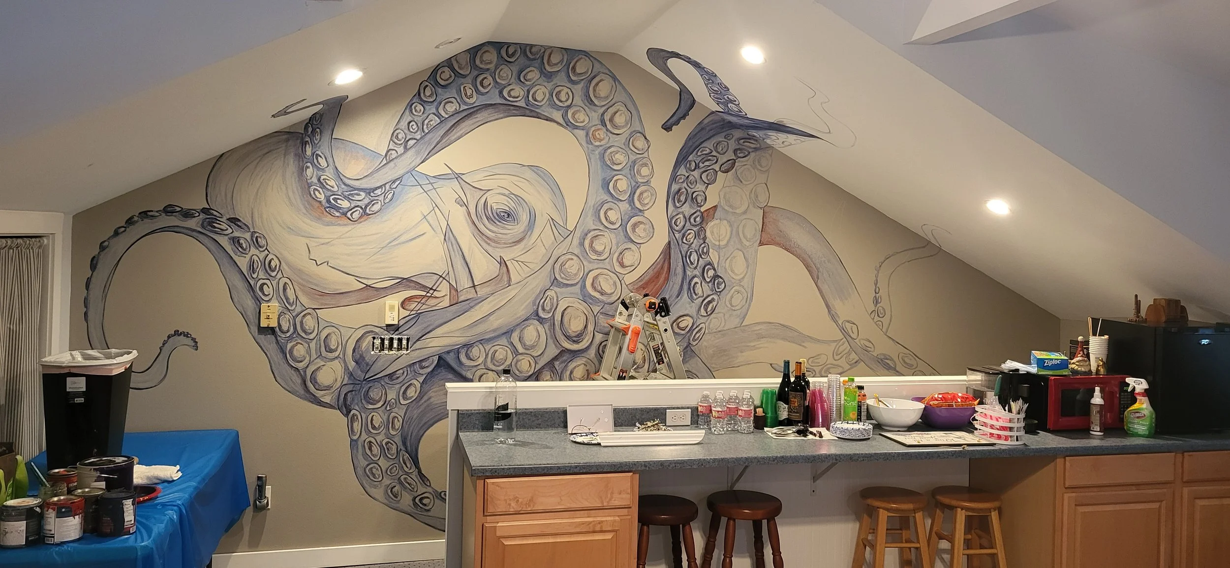 A mural of an octopus on a wall above a kitchen counter, with bottles, bowls, cups, and a microwave on the counter, and stools in front.