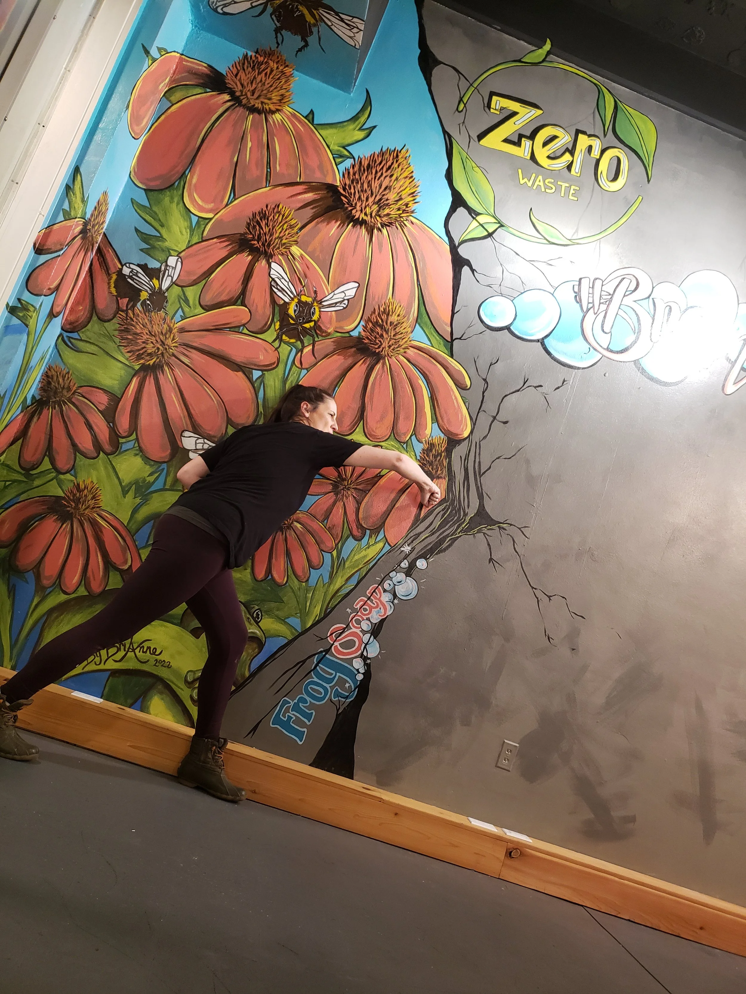 A woman is painting a colorful mural of flowers, bees, and branch on a wall. The mural includes text that reads 'Zero Waste' and 'Trash.'