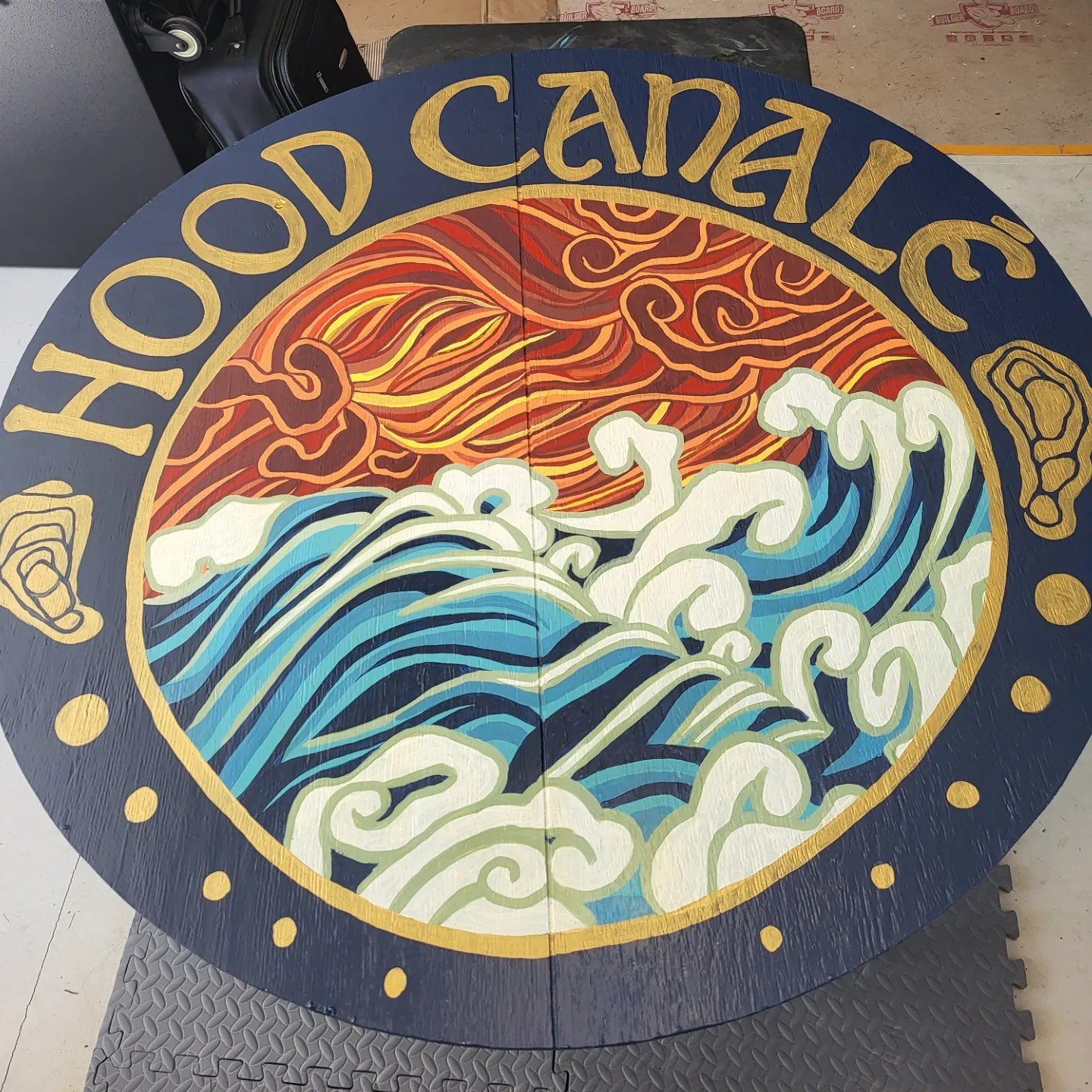 A round wooden sign with the words 'Hot Food Caddyale' written around the top edge in gold. The center features a colorful, stylized depiction of a wave with white froth, a sun with swirling red and orange rays, and Japanese-style clouds, all painted