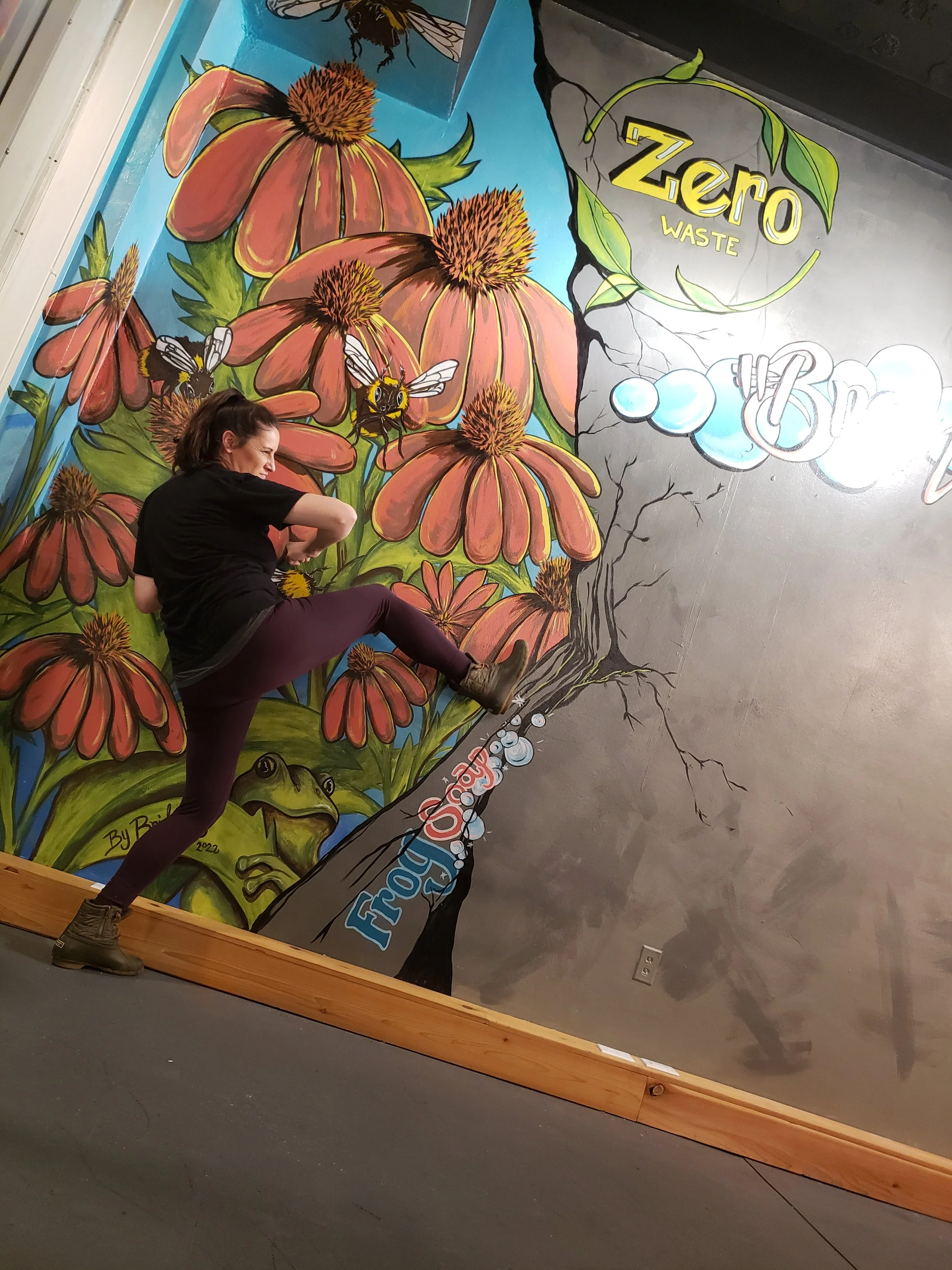 A woman standing on a wooden stage in front of a colorful mural featuring large pink flowers, bees, frogs, and words related to waste reduction, with the word "Zero" in green and yellow above.