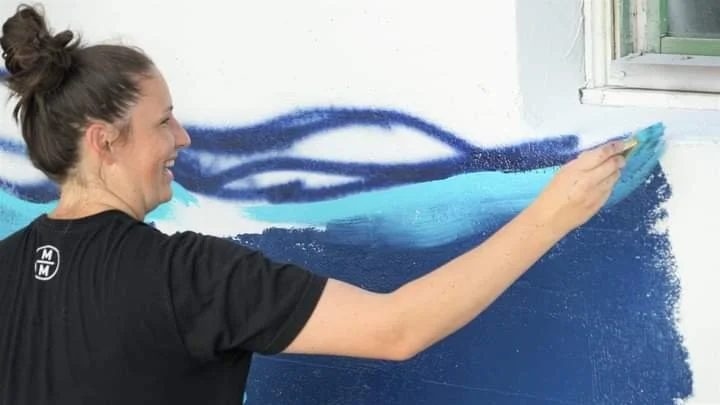 A woman painting a mural on a white wall, with blue shades, near a window.