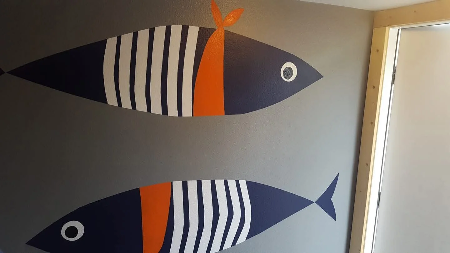 Decorative wall with two stylized fish illustrations, painted in navy blue, orange, and white, hanging on a light gray wall near a wooden door frame.