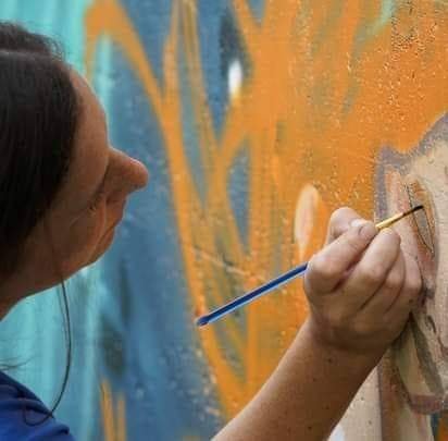 A woman painting a colorful mural on a wall with a small paintbrush.
