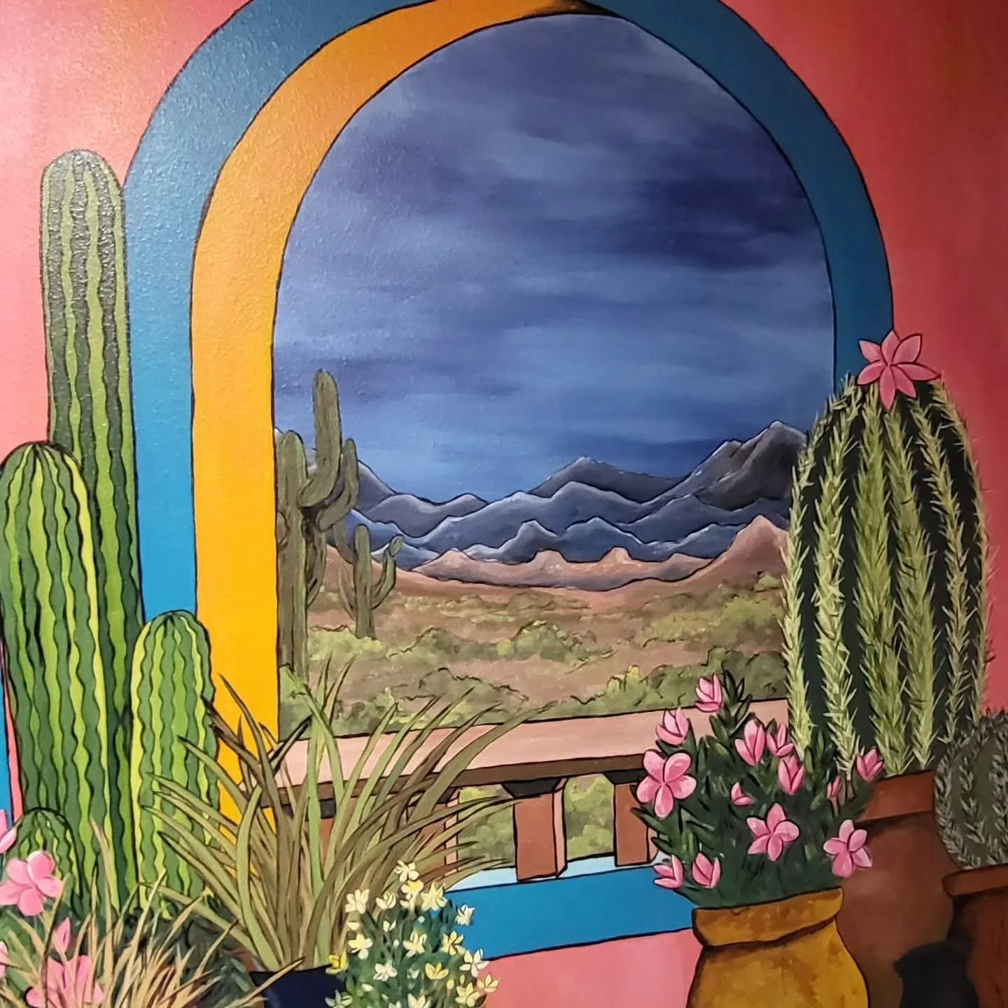 Colorful indoor mural of a desert landscape with cacti, mountains, and a cloudy sky, framed by a rainbow-shaped arch and decorated with potted pink flowers.