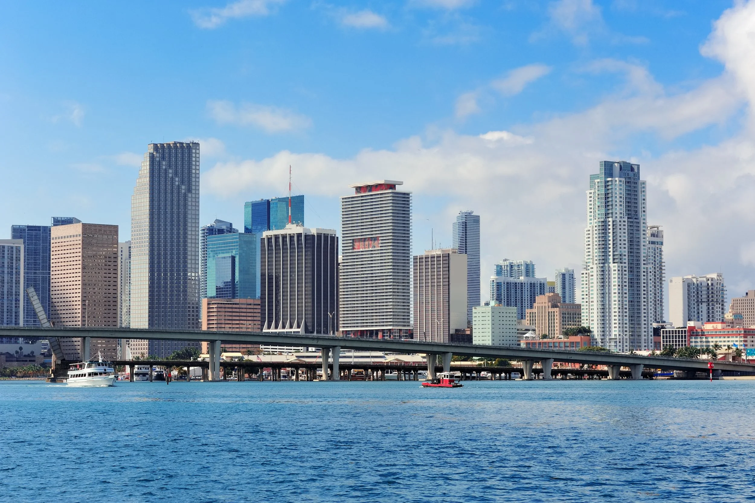 City skyline with tall modern skyscrapers viewed from the water under a blue sky with scattered clouds, with boats on the water and a bridge in the foreground.