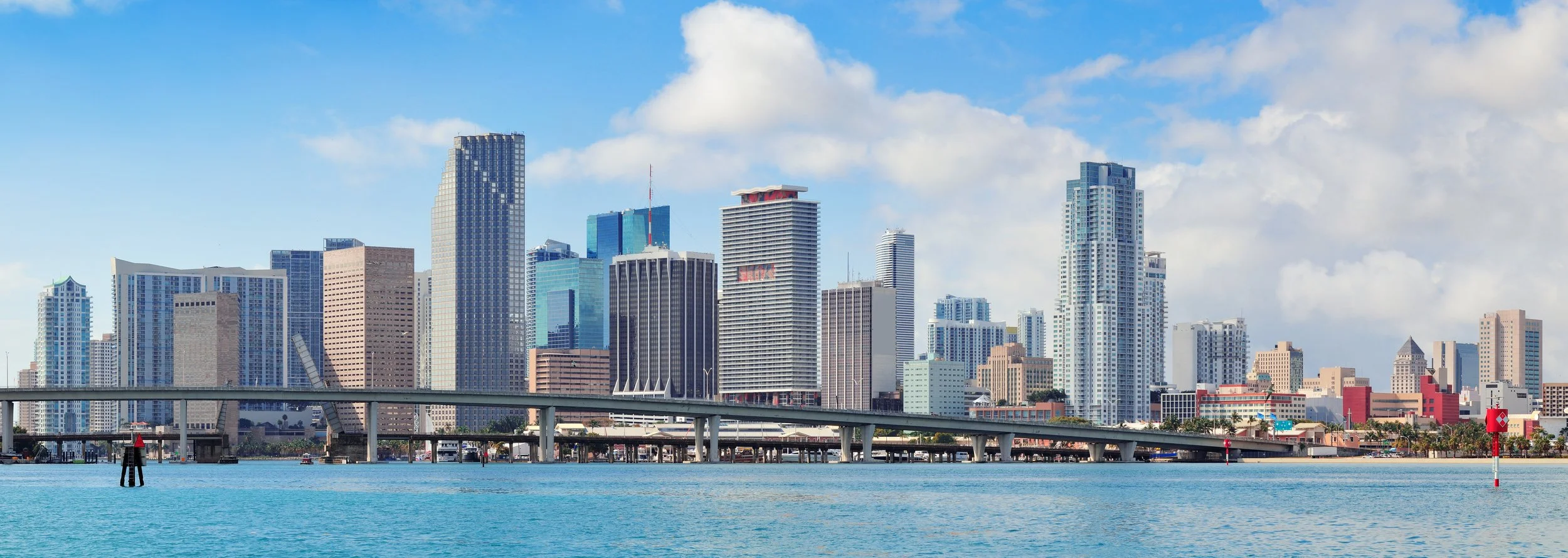 Skyline of downtown Miami with various tall modern buildings, a bridge over turquoise water, and partly cloudy blue sky.
