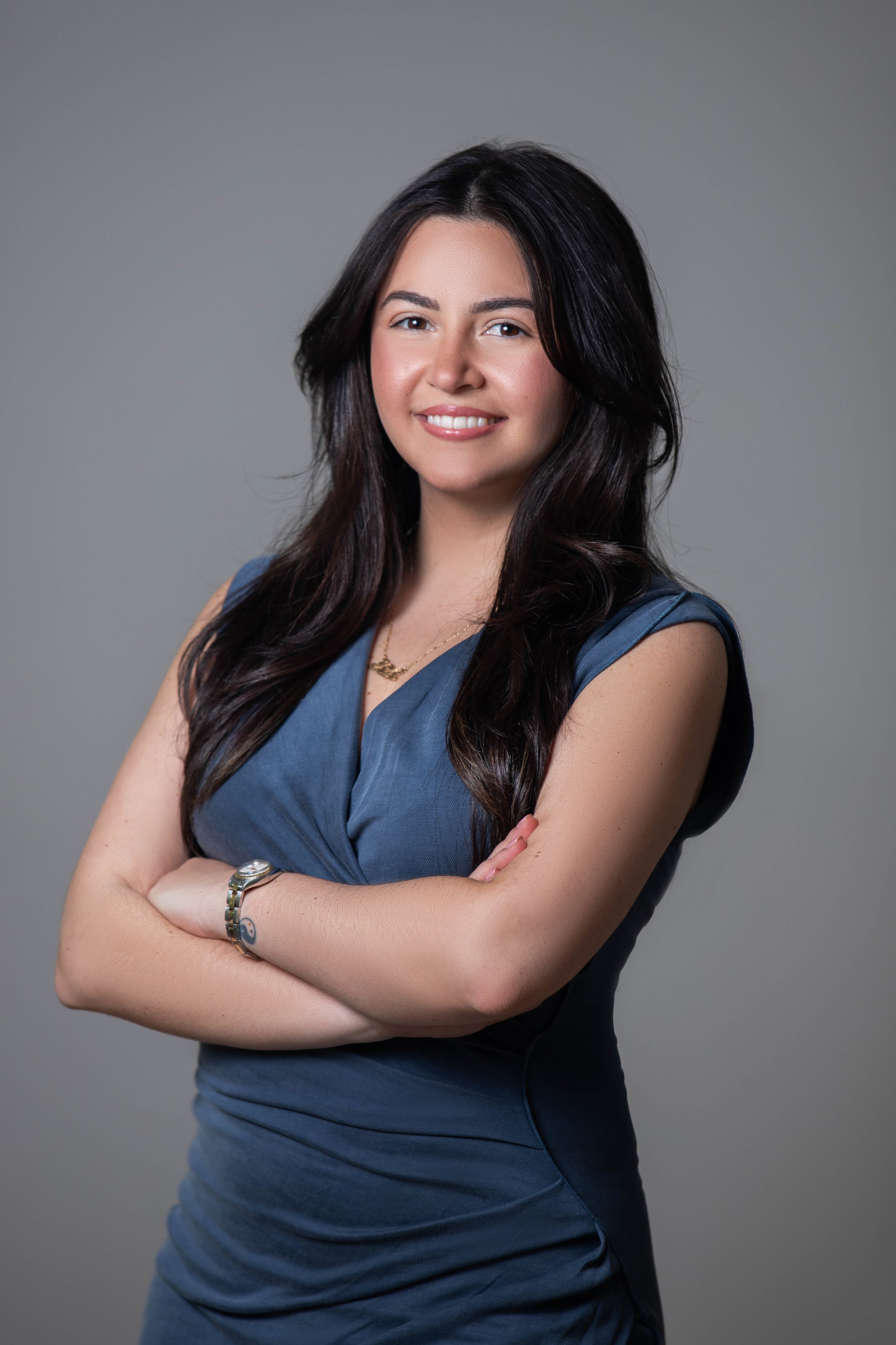 A young woman with long dark hair smiling, wearing a sleeveless dark blue dress, standing with crossed arms against a gray background.