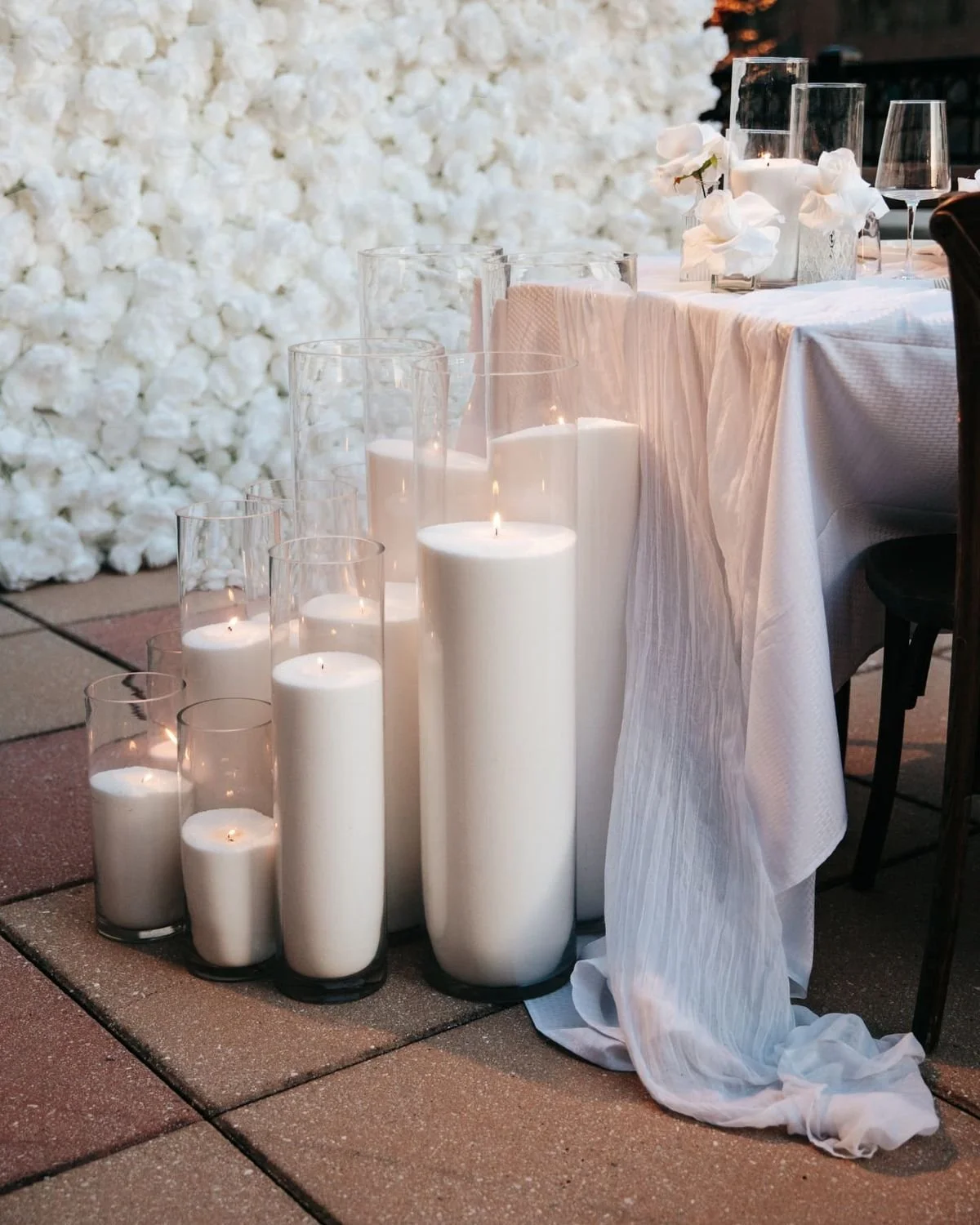A table decorated with white pearl wax candles of varying heights in glass holders, white flowers in a vase, and a white tablecloth, set against a background of white floral decorations, likely for a wedding or an elegant event.