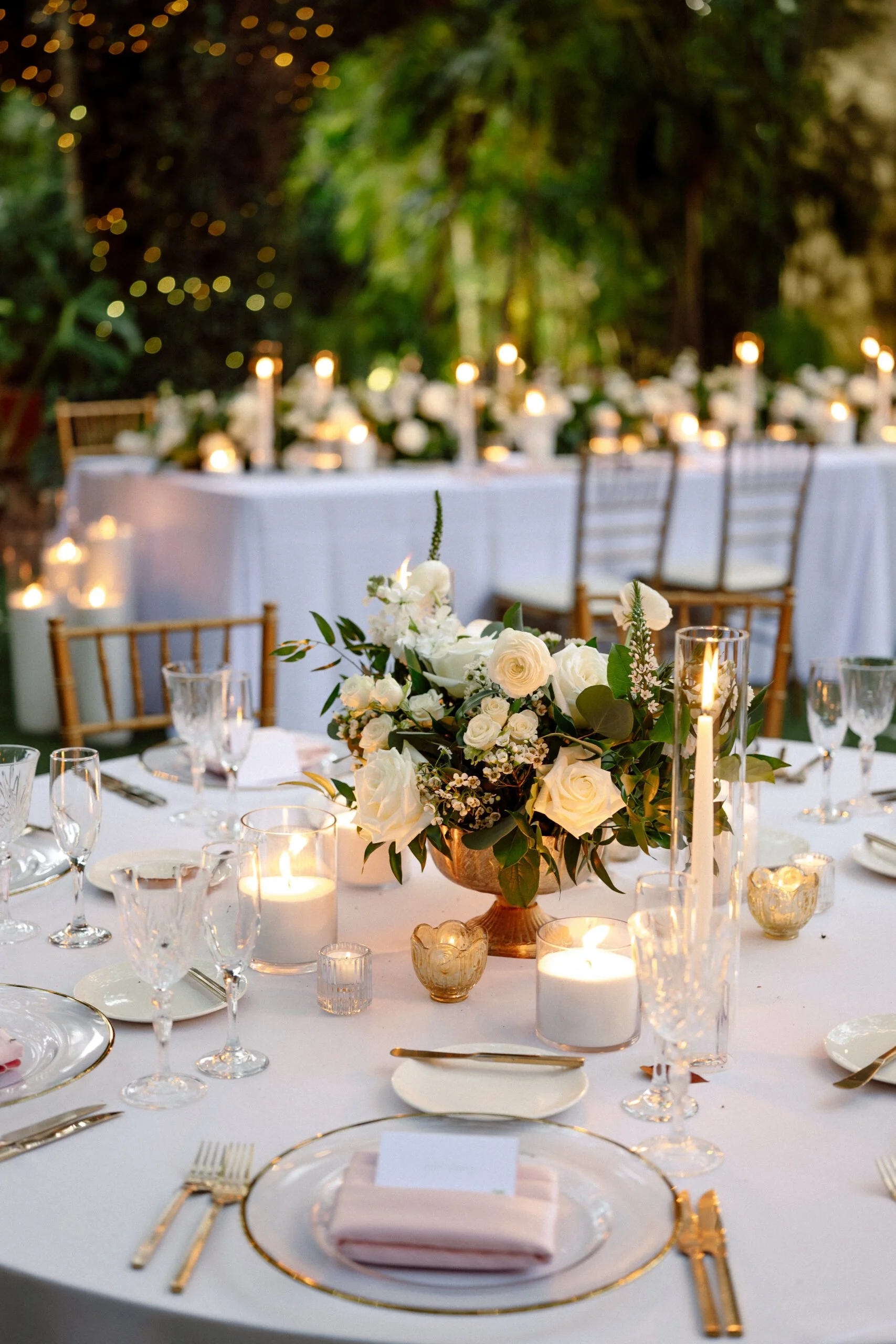 Elegant outdoor wedding reception table decorated with white flowers, pearl wax candles, and gold-rimmed glassware and cutlery, set in a lush green garden with string lights.