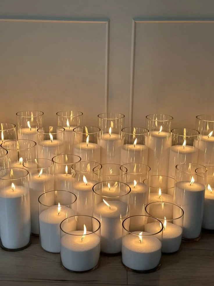 Several lit white pearl wax candles in glass holders arranged on a light-colored surface in front of a white wall with paneling.