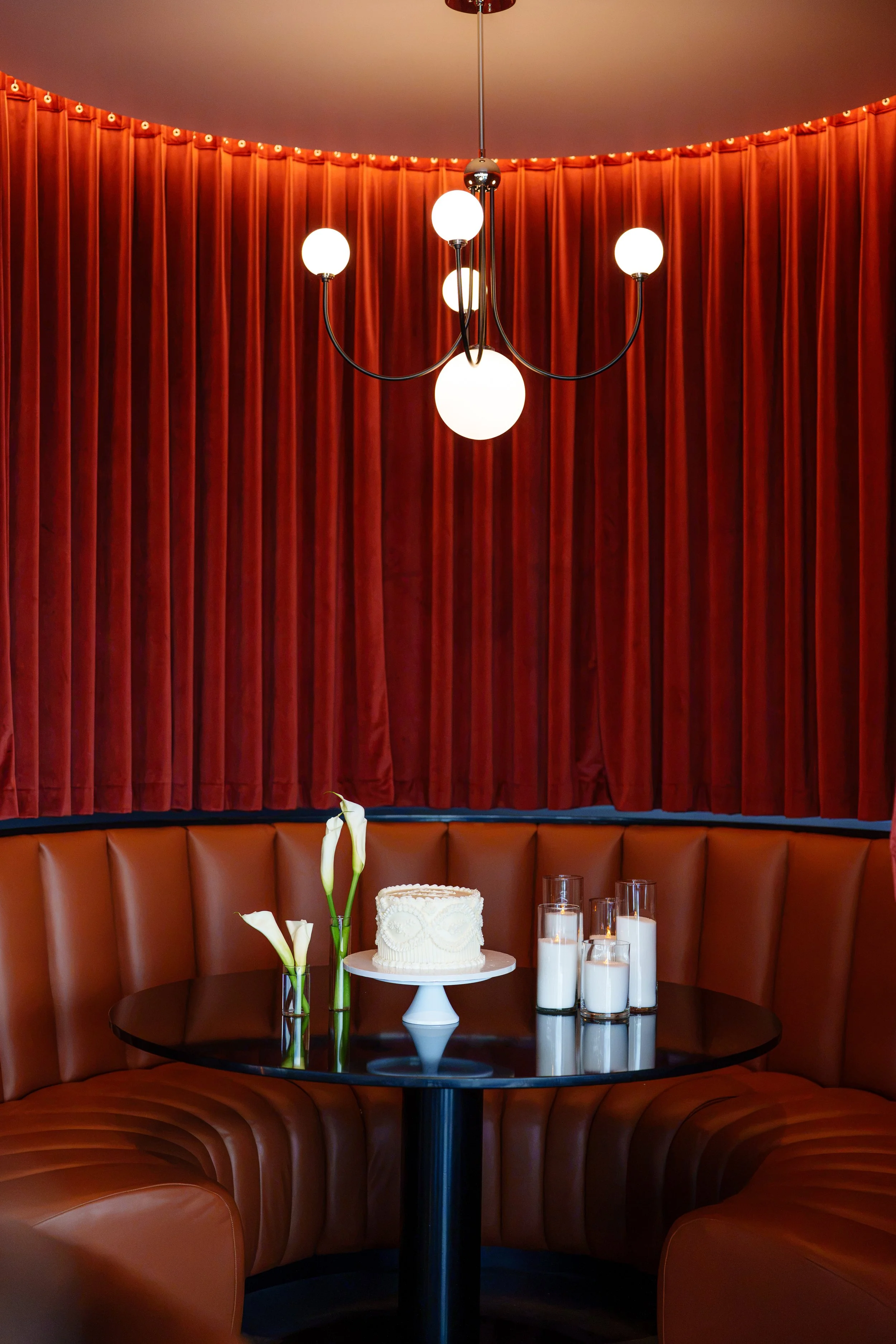 Intimate lounge setting with a white wedding cake, white calla lilies, and glowing Pearl & Ember pearl wax candles against rich red velvet draping and cognac leather seating. Photography: Nicole Dixon Photographic