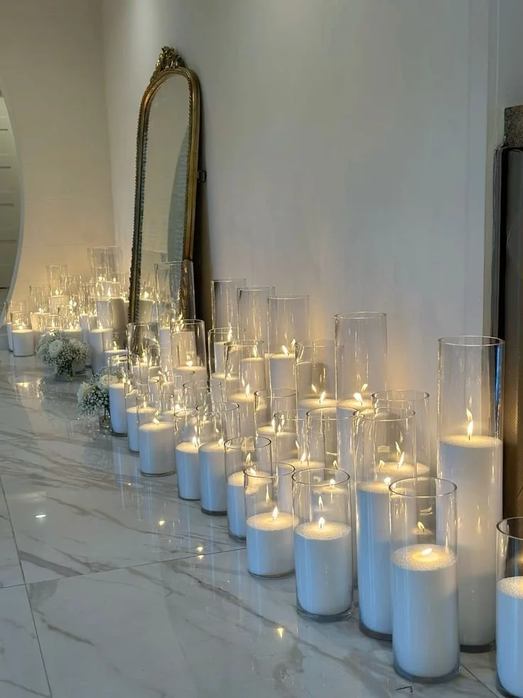 A row of white pearl wax candles in tall glass vessels with lit flames, placed along a marble surface, with a large gold-framed mirror leaning against the wall and some small floral arrangements nearby.