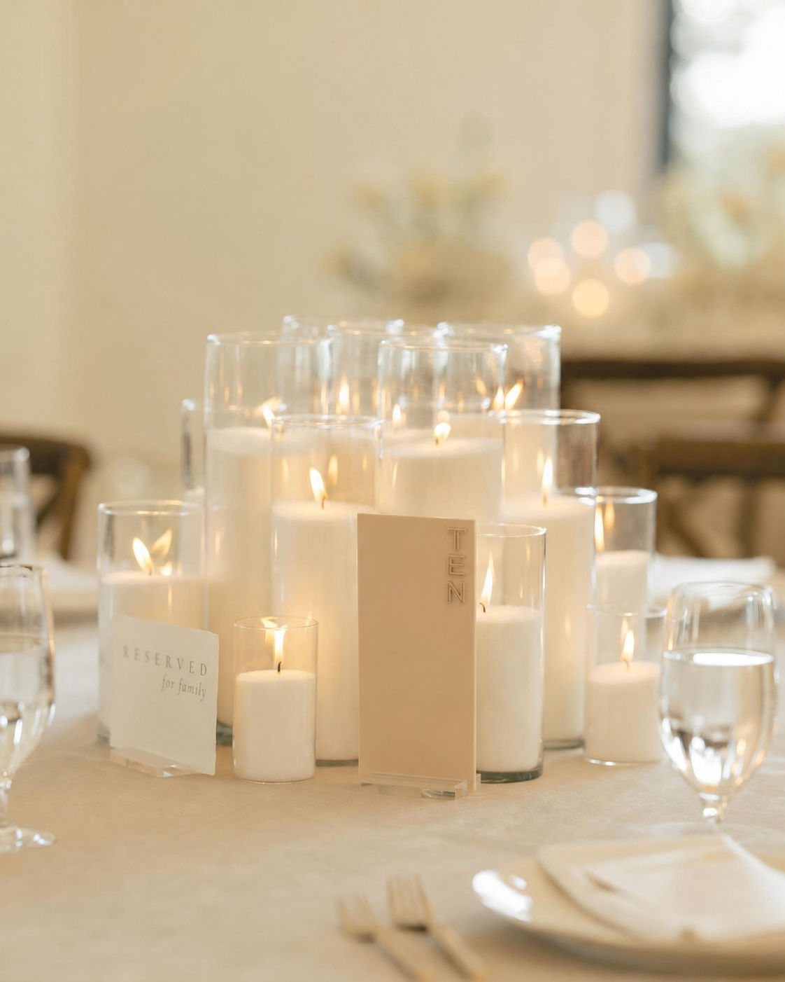 Table centerpiece with multiple lit white pearl wax candles in glass holders, reserved sign, and tent card at event.