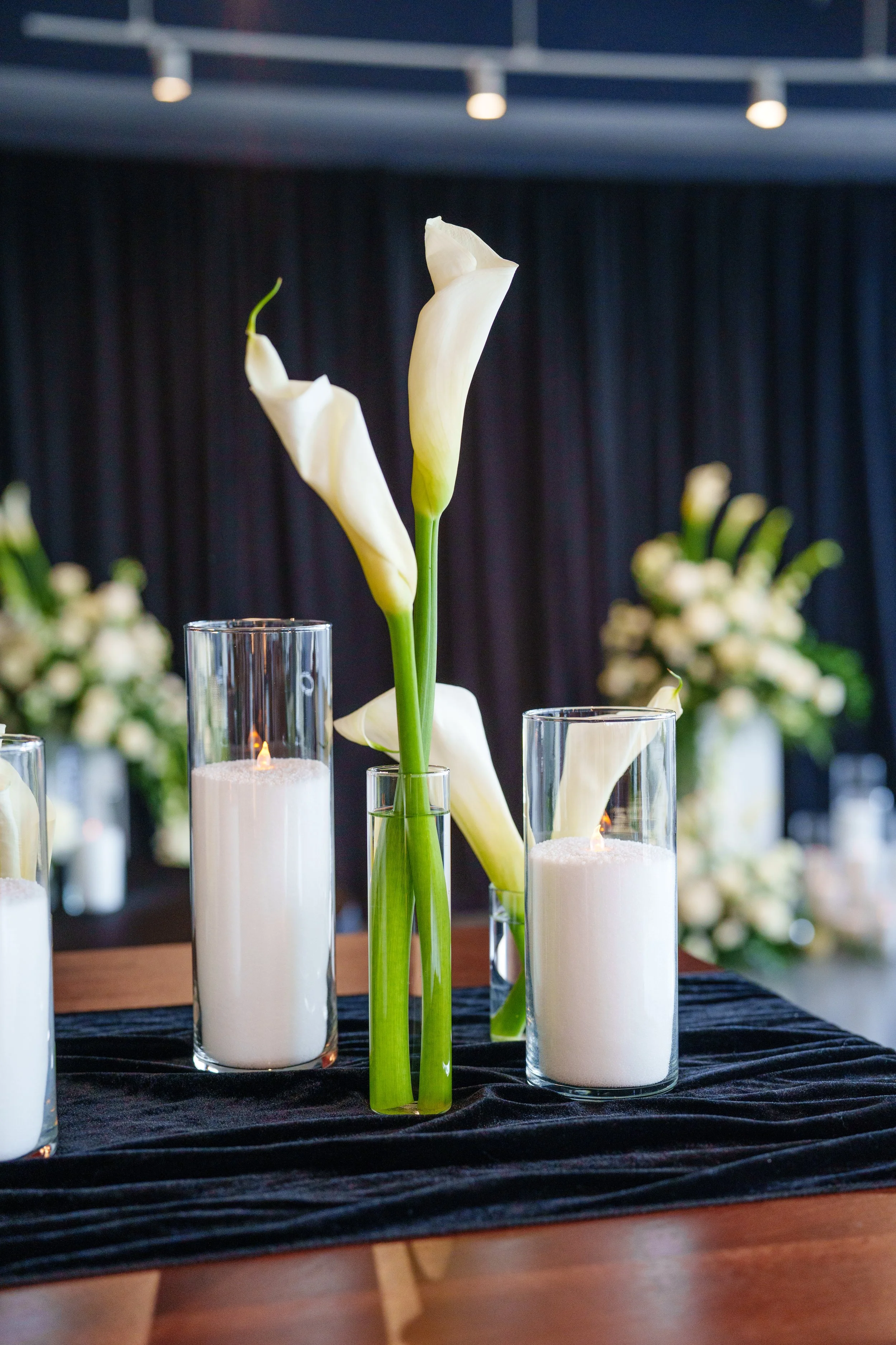 Close-up of Pearl & Ember pearl wax candles in glass vessels paired with elegant white calla lilies on a dark velvet table runner, creating a sophisticated candlelit event setting. Photography: Nicole Dixon Photographic