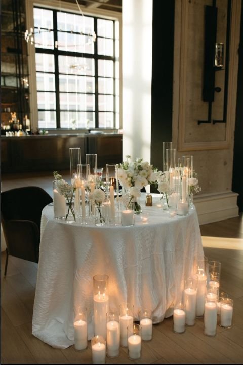 Elegant table decorated with white flowers and multiple pearl wax candles in glass holders, set in a warmly lit room with large windows.