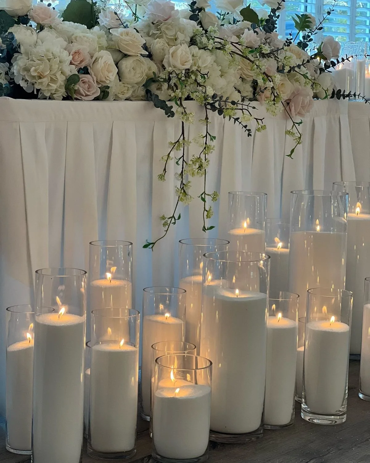 A floral arrangement with white and blush roses, white hydrangeas, and greenery, decorated with burning pearl wax candles in glass holders on a table with white drapery.