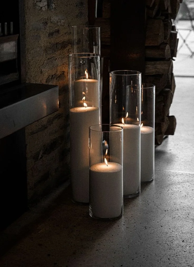 Four tall glass cylinders contain white pearl wax candles with flames, placed on a concrete floor near a stone wall and a stack of wood.