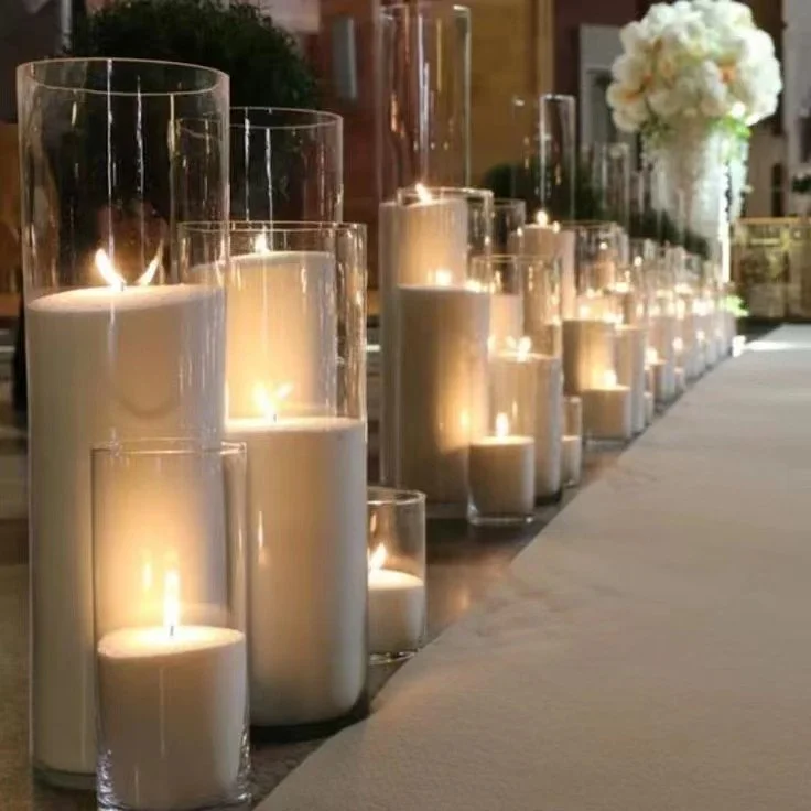 A row of lit white pearl wax candles in tall glass vases, with a floral arrangement in the background, for a wedding or special event.