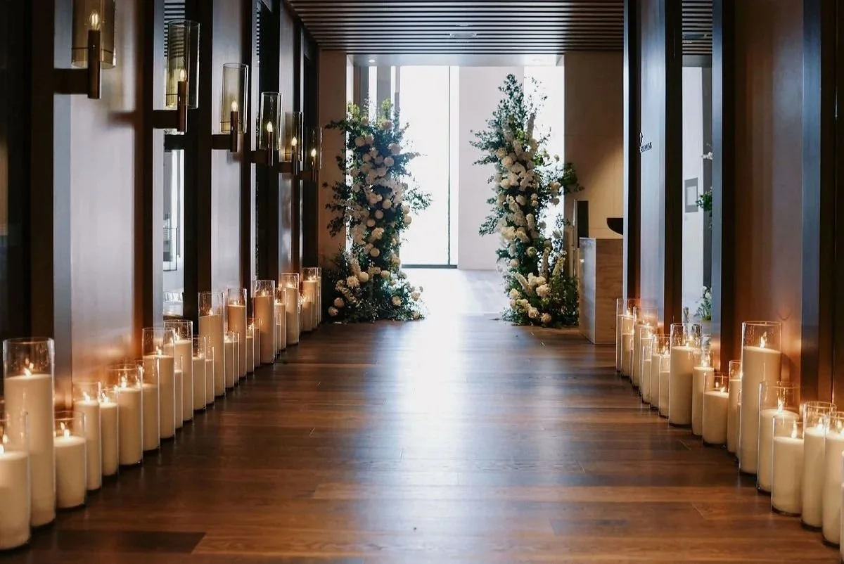 Elegant hallway decorated for an event with tall flower arrangements and pearl wax candles lining the floor.