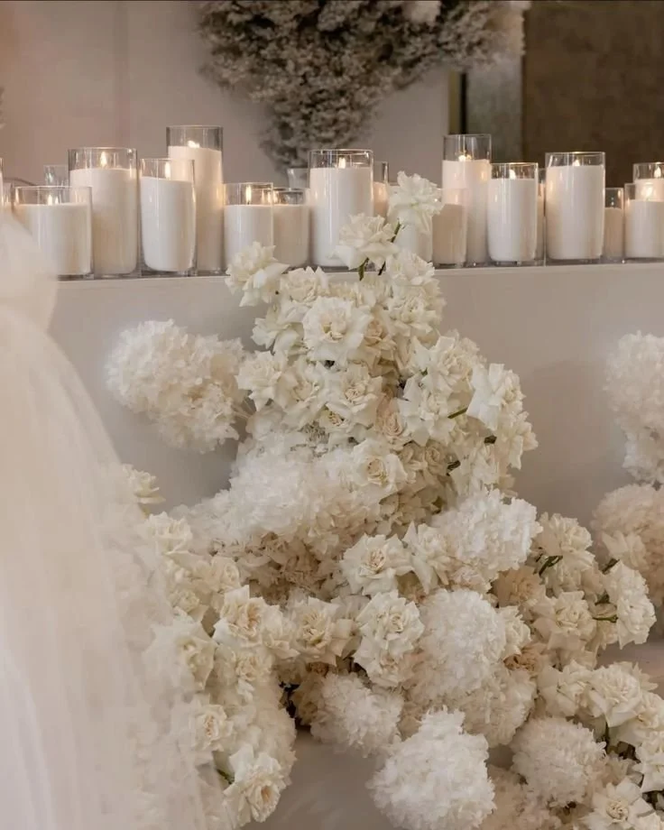 A collection of white pearl wax candles in glass holders and a large floral arrangement of white flowers on a table.