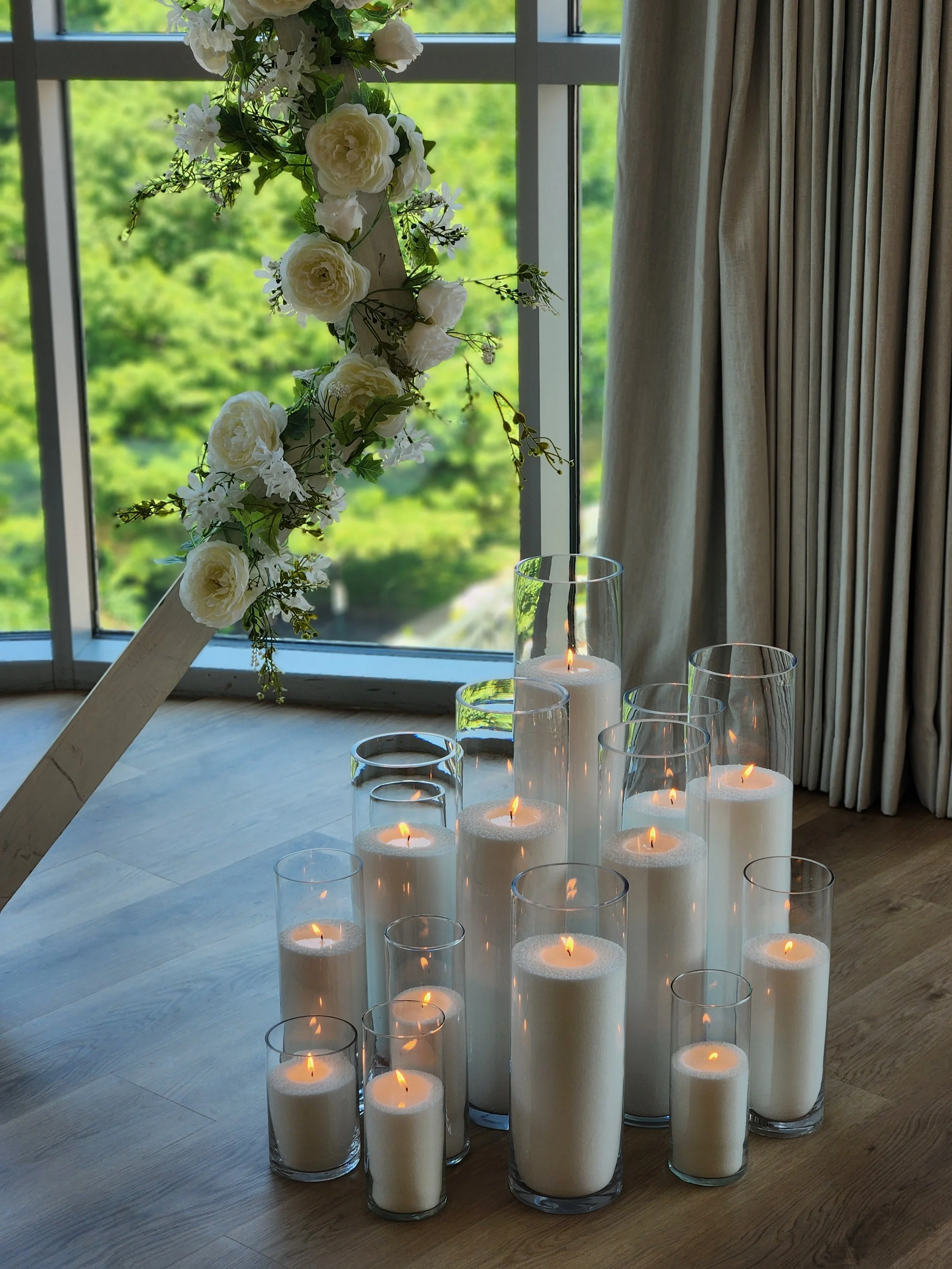 Luxury pearl wax candles in clear glass vessels arranged in a large floor cluster beside a white floral wedding installation at a Columbus, Ohio event venue.
