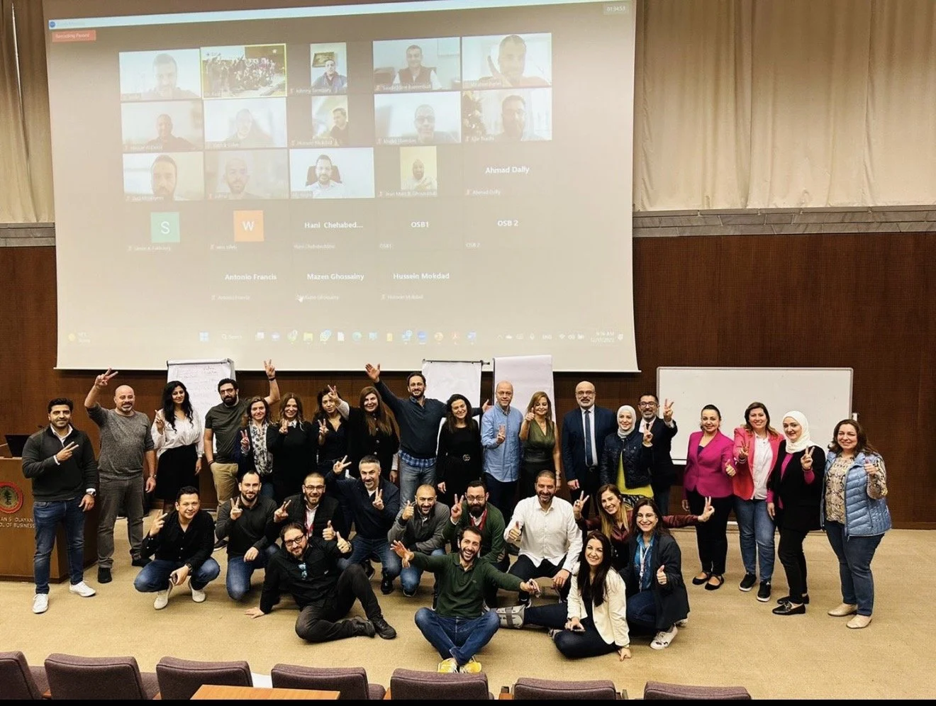 A large group of diverse people posing for a photo in a conference room with a projector screen showing a video call behind them.