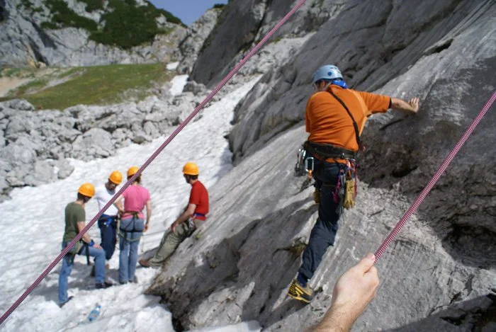 Group of people climbing a rocky mountain with safety ropes, wearing helmets and harnesses, surrounded by snow and mountains.