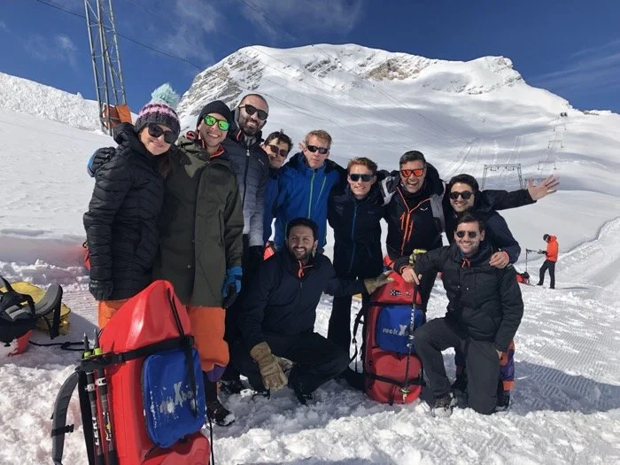 Group of nine people dressed in winter gear posing for a photo in snowy mountain landscape, with peaks and a clear blue sky in the background.