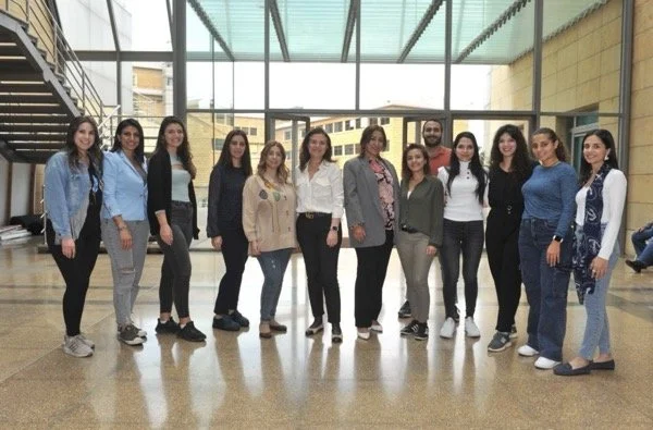 Group of 12 women standing indoors, posing for a photo in a bright, modern building with large windows and stairs.