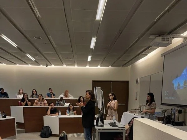 A group of people attending a lecture or presentation in a classroom or conference room. A woman is standing at the front, speaking to the audience, with a presentation slide projected on the wall.
