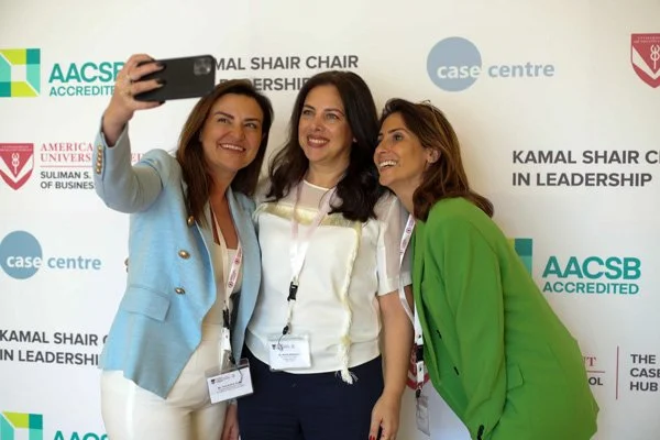 Three women taking a selfie in front of a backdrop with logos for Kamal Shair School of Leadership, AACSB Accredited, and Case Centre. The woman on the left is wearing a light blue blazer, the middle woman is wearing a white top, and the woman on the