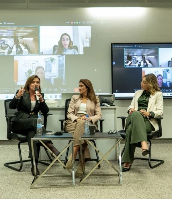 Three women seated in a panel discussion in a professional setting, with two large screens displaying a virtual meeting behind them.