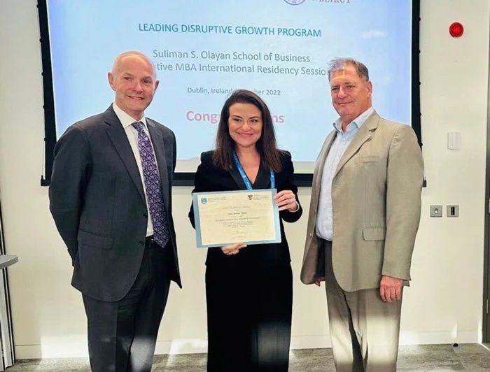Woman holding a diploma flanked by two men in business suits in a classroom or conference room, with a presentation screen behind them displaying text related to a business program at Suliman S. Olayan School of Business in Dublin, Ireland.