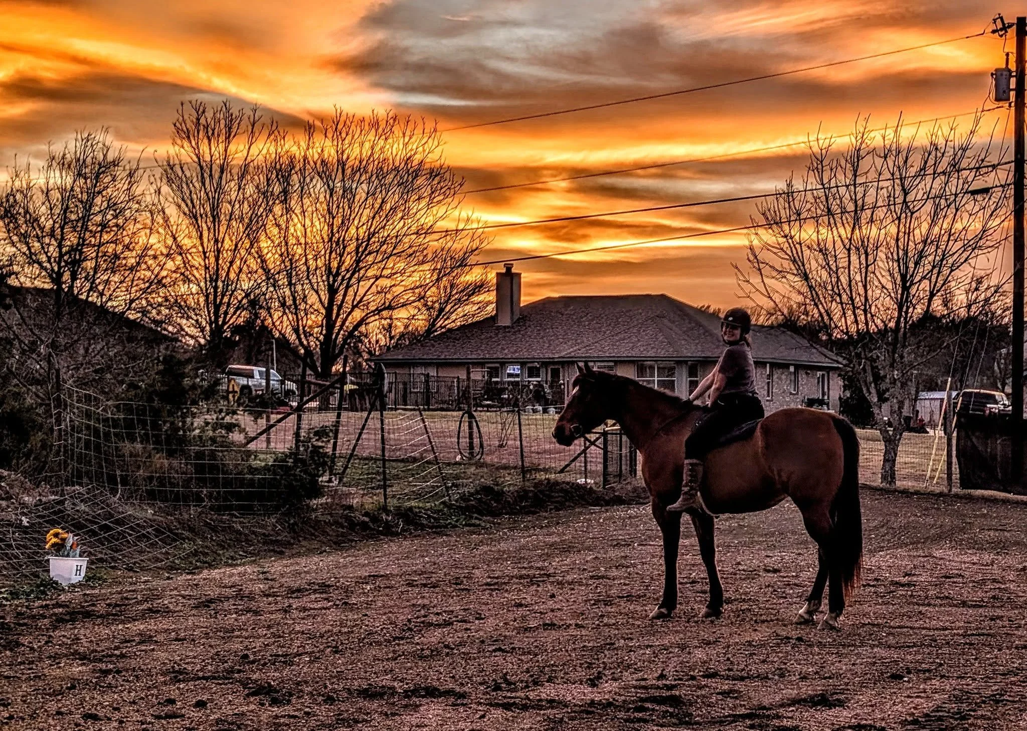 A person riding a horse in an outdoor area during sunset, with a house, leafless trees, power lines, and various objects in the background.