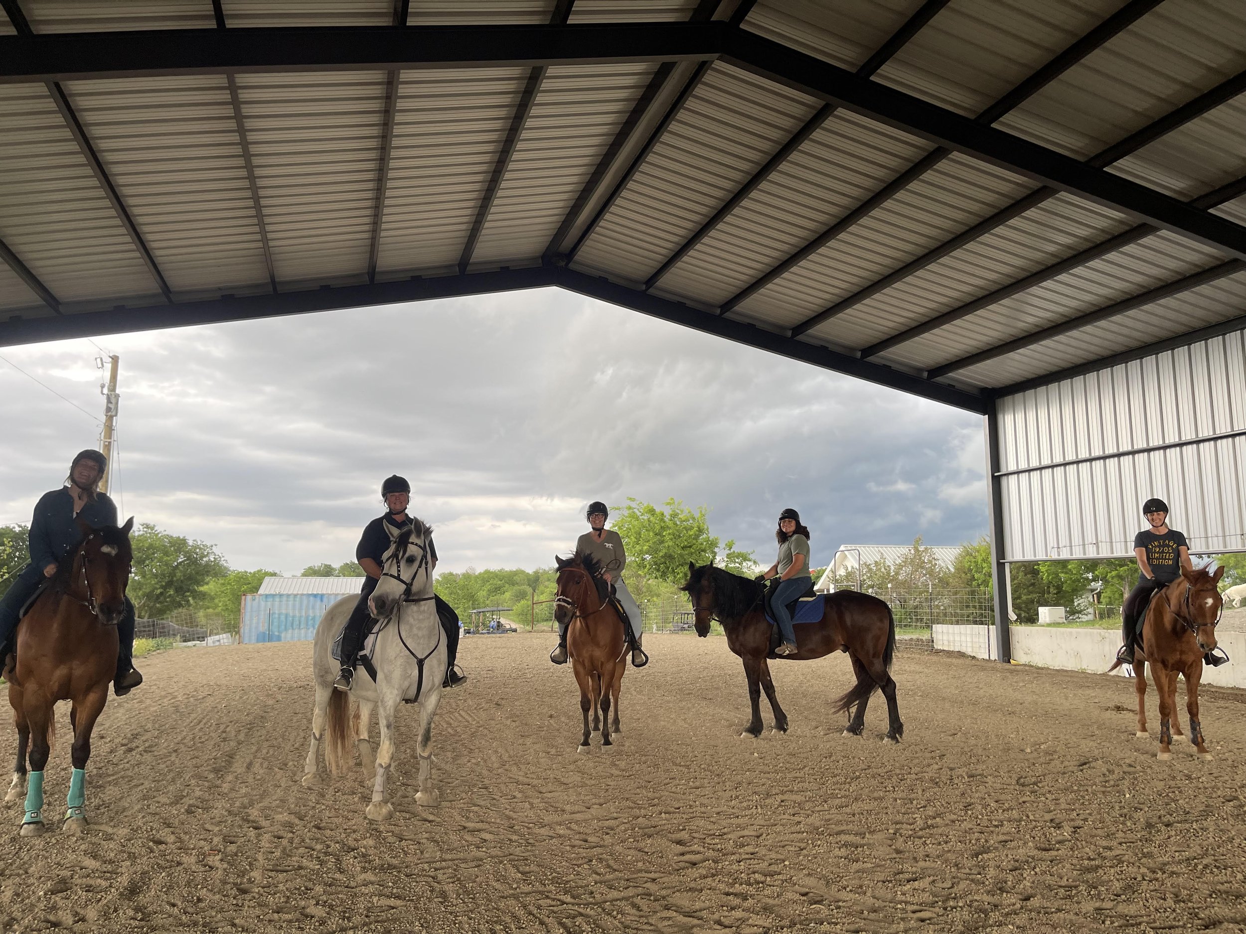 Group of people riding horses in an indoor riding arena with a metal roof and a cloudy sky outside.