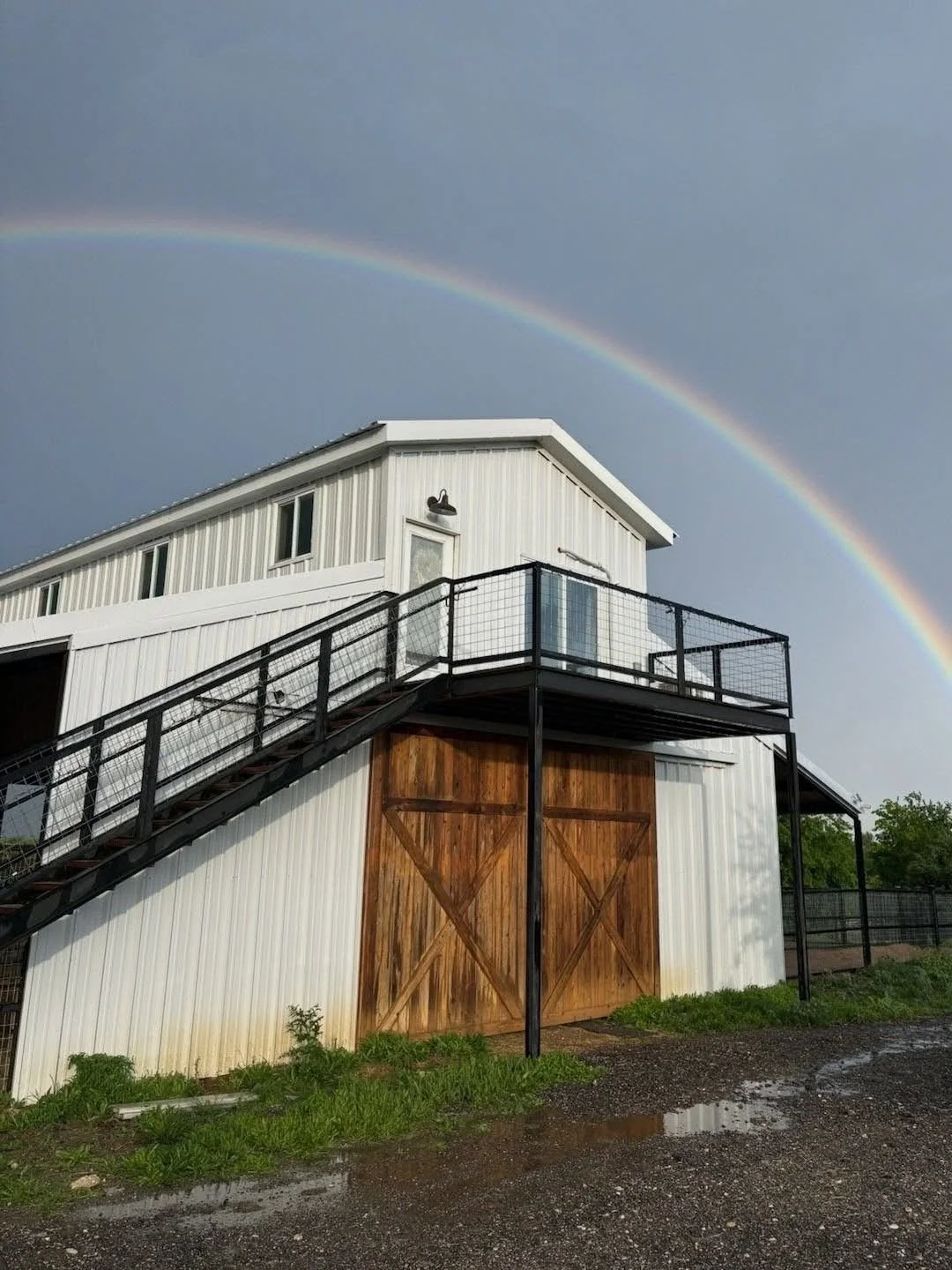 A white barn-style building with a wooden sliding door, surrounded by grass and a gravel path, under a sky with a rainbow.