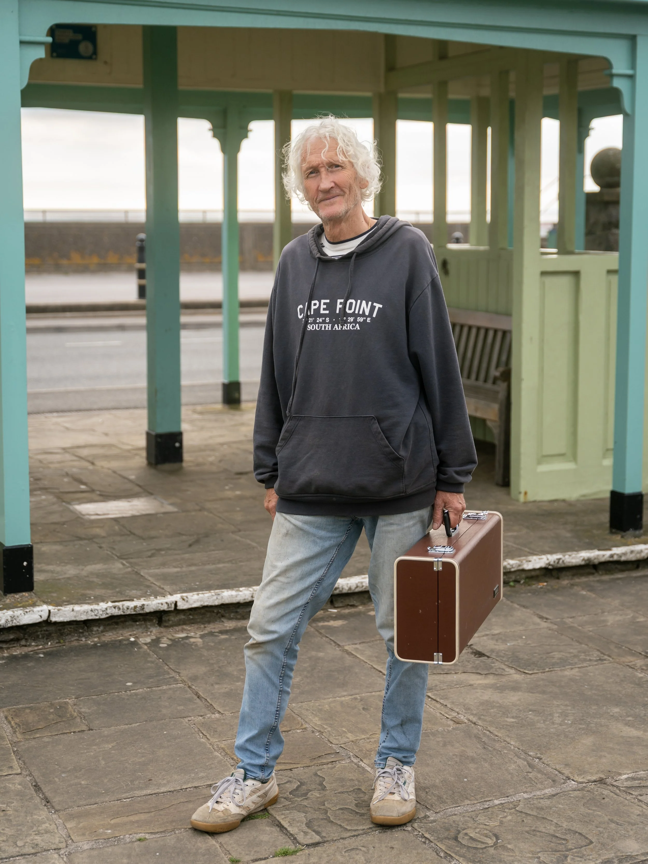 A man standing at a bus stop with a briefcase in his right hand, wearing a black hoodie and light jeans.