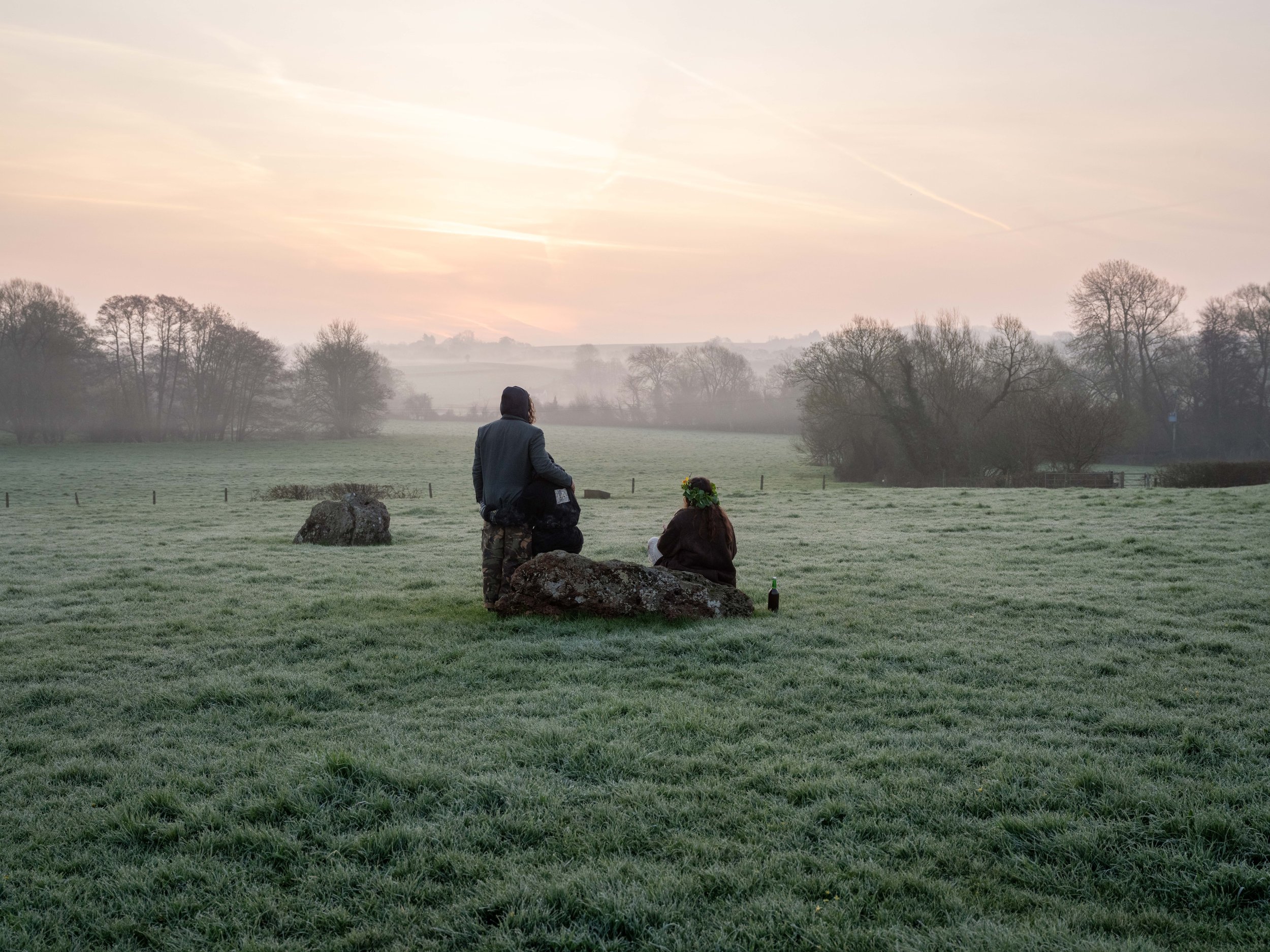 Two people in a grassy field at sunrise; one person standing, the other sitting on a rock, with a bottle nearby, surrounded by trees and mist.