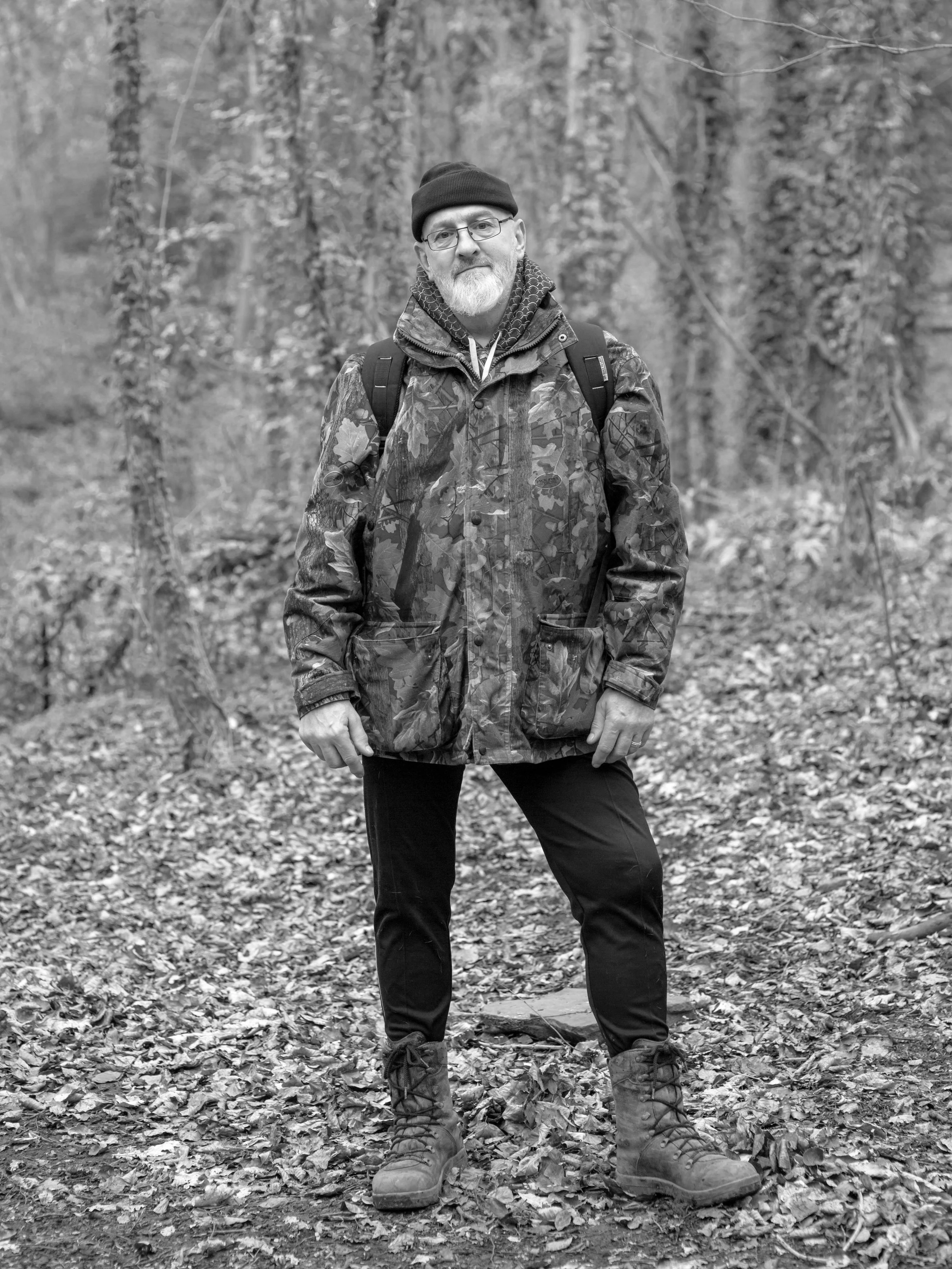 A man standing outdoors on a wooded trail, wearing outdoor gear including a camouflage jacket, black pants, hiking boots, a dark beanie, glasses, and carrying a backpack.