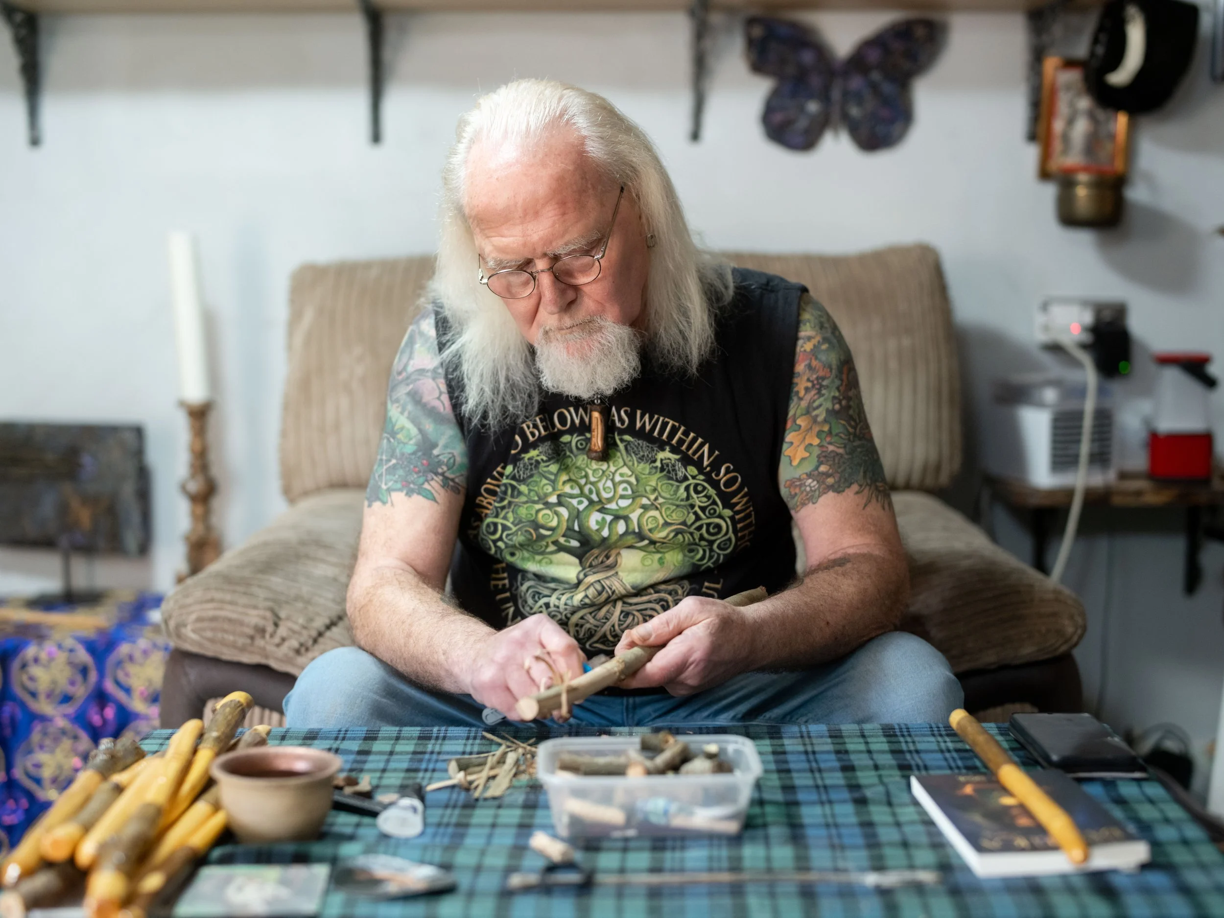 An elderly man with long white hair and a beard, wearing glasses and a black T-shirt with a green tree design, is sitting at a table working on a woodcraft project. The table is covered with a green and blue plaid cloth and has various wooden tools a