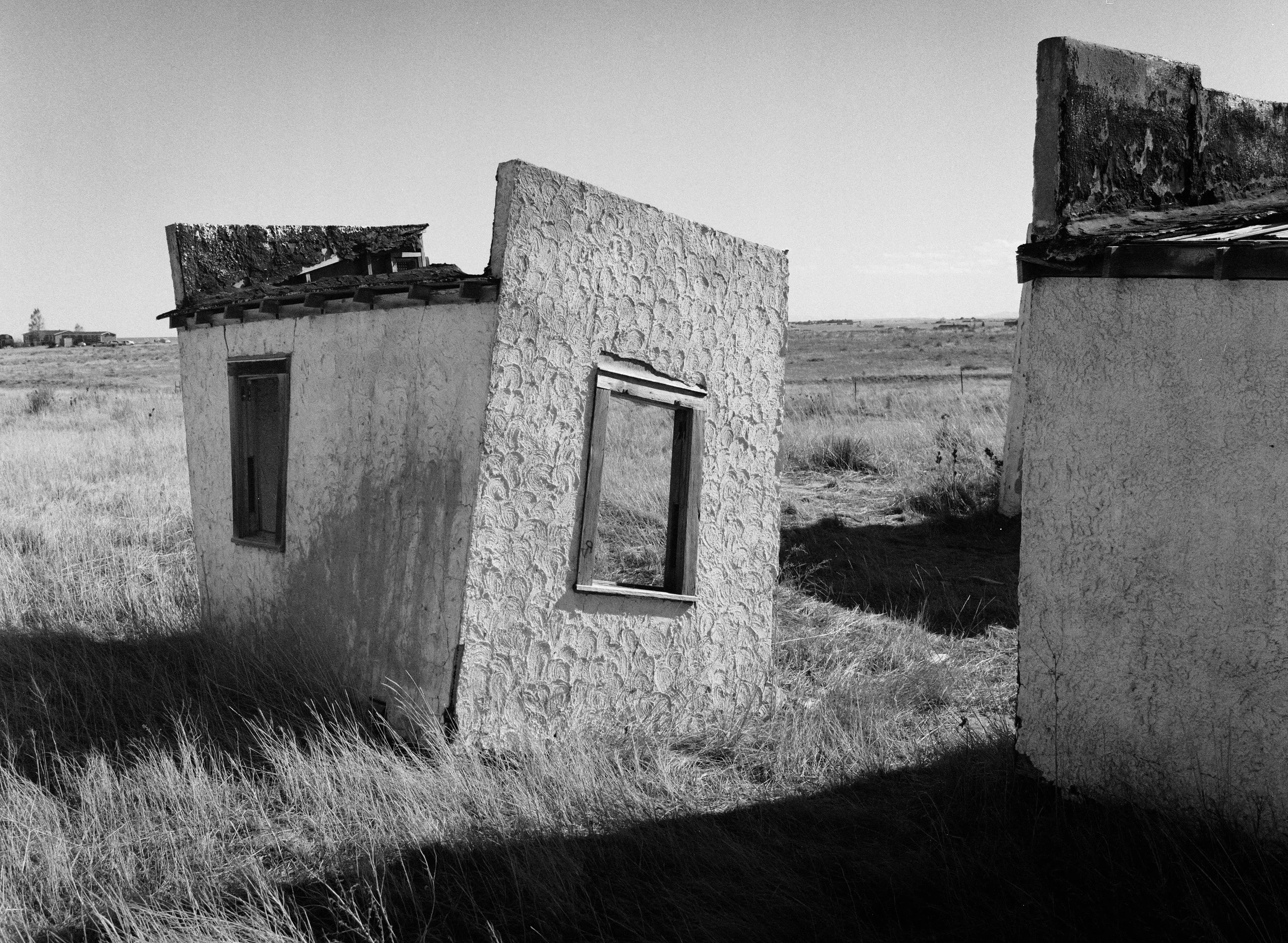 Abandoned, tilted small building with textured exterior walls and missing or damaged roof, located in a field with tall grass, under a clear sky.