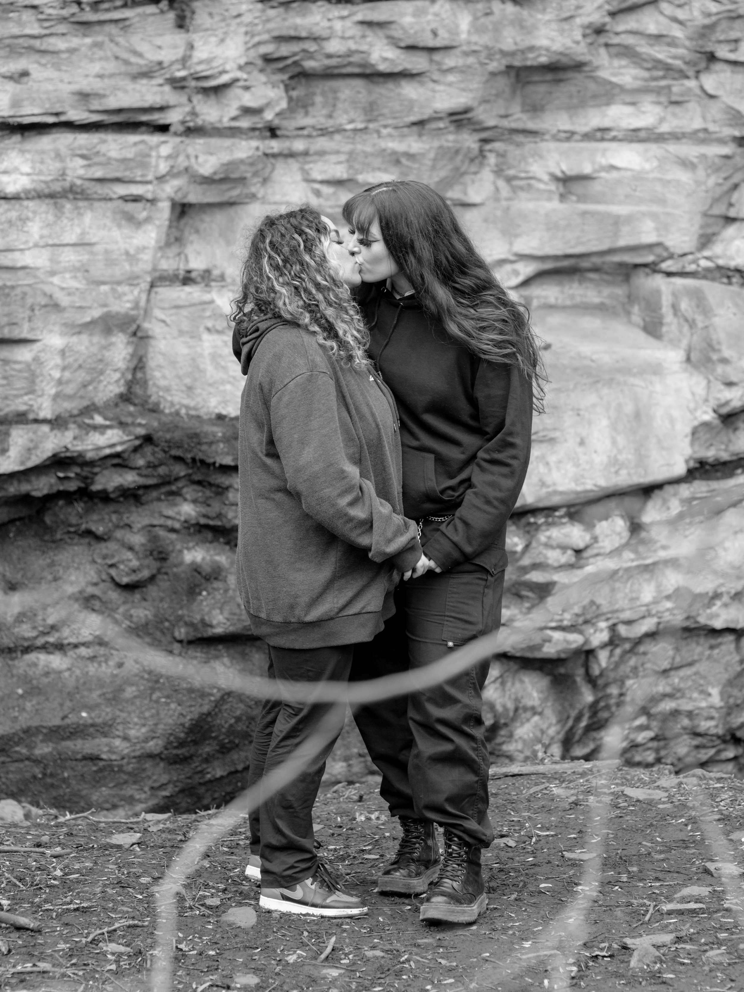Two women sharing a kiss while holding hands outdoors in front of a rocky background.
