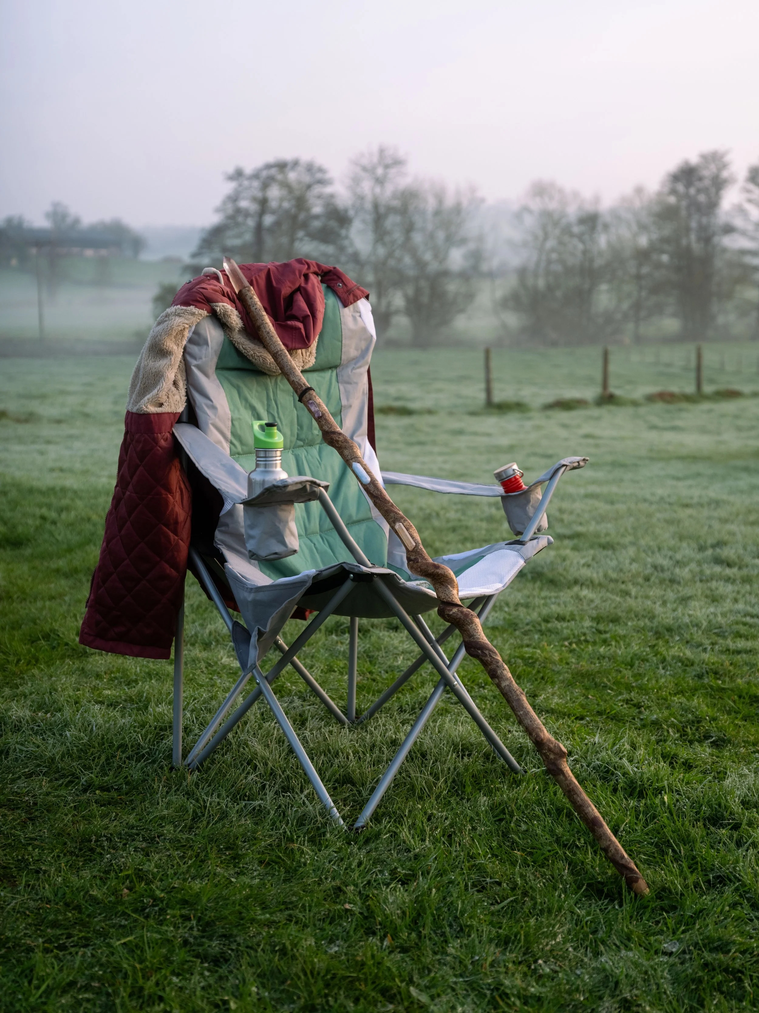 An empty outdoor camping chair with a coat draped over it, a walking stick leaning against it, and a water bottle attached to its armrest, set on a grassy field with trees in the background.