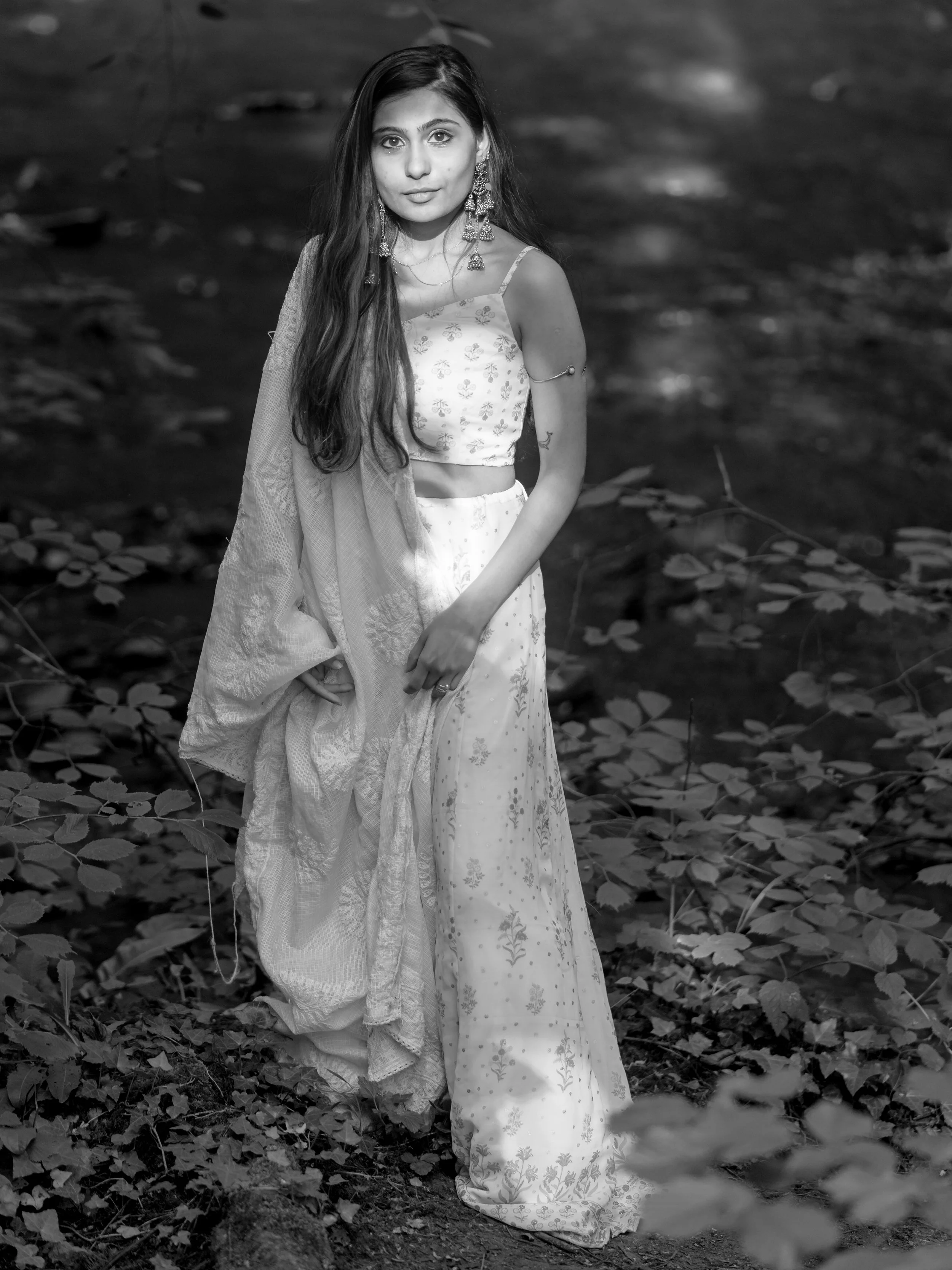 A young woman wearing traditional Indian attire, standing outdoors among foliage, looking at the camera.