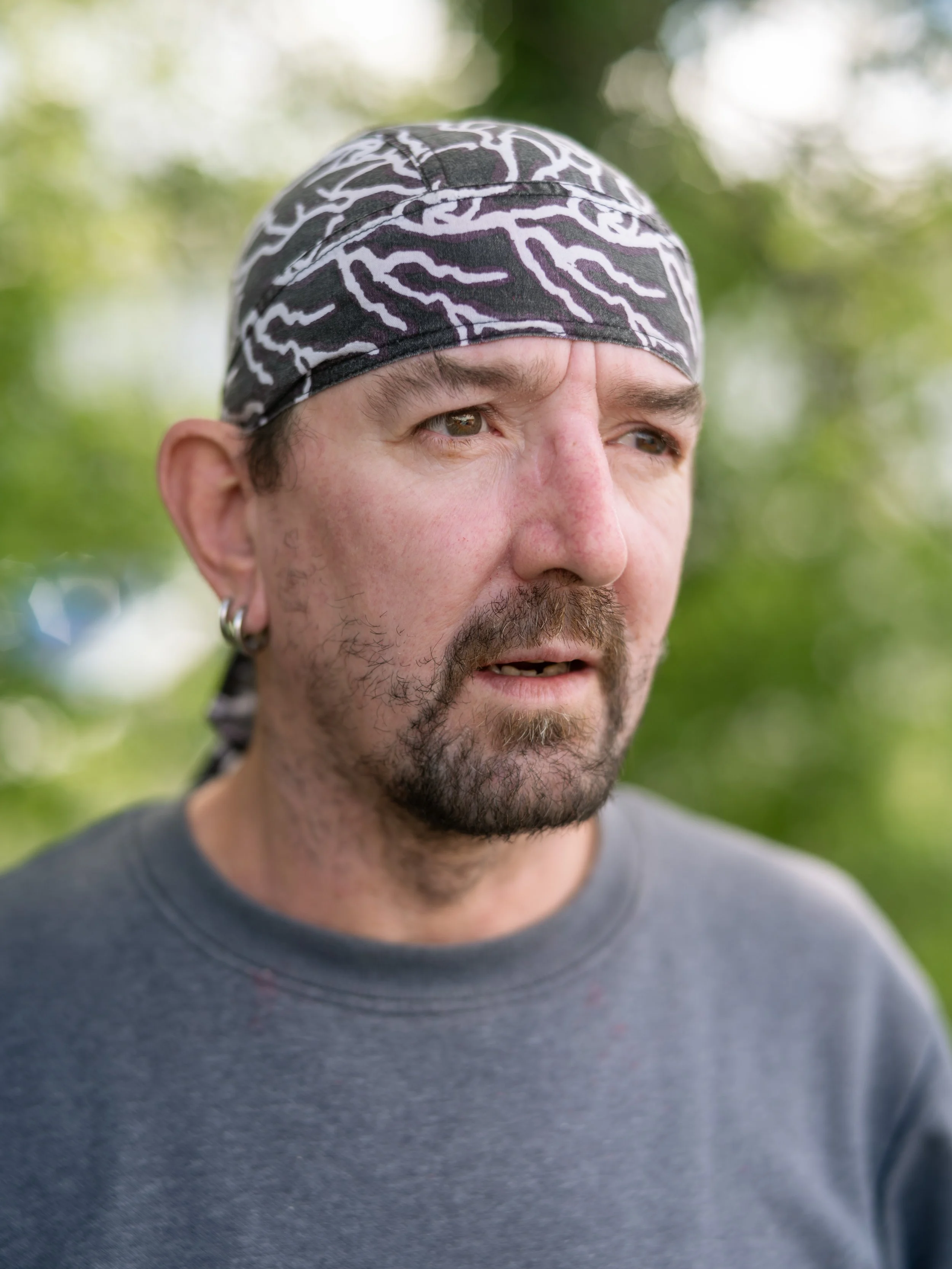 A man with a bandana on his head and an earring in his left ear is outdoors with a blurred green background.