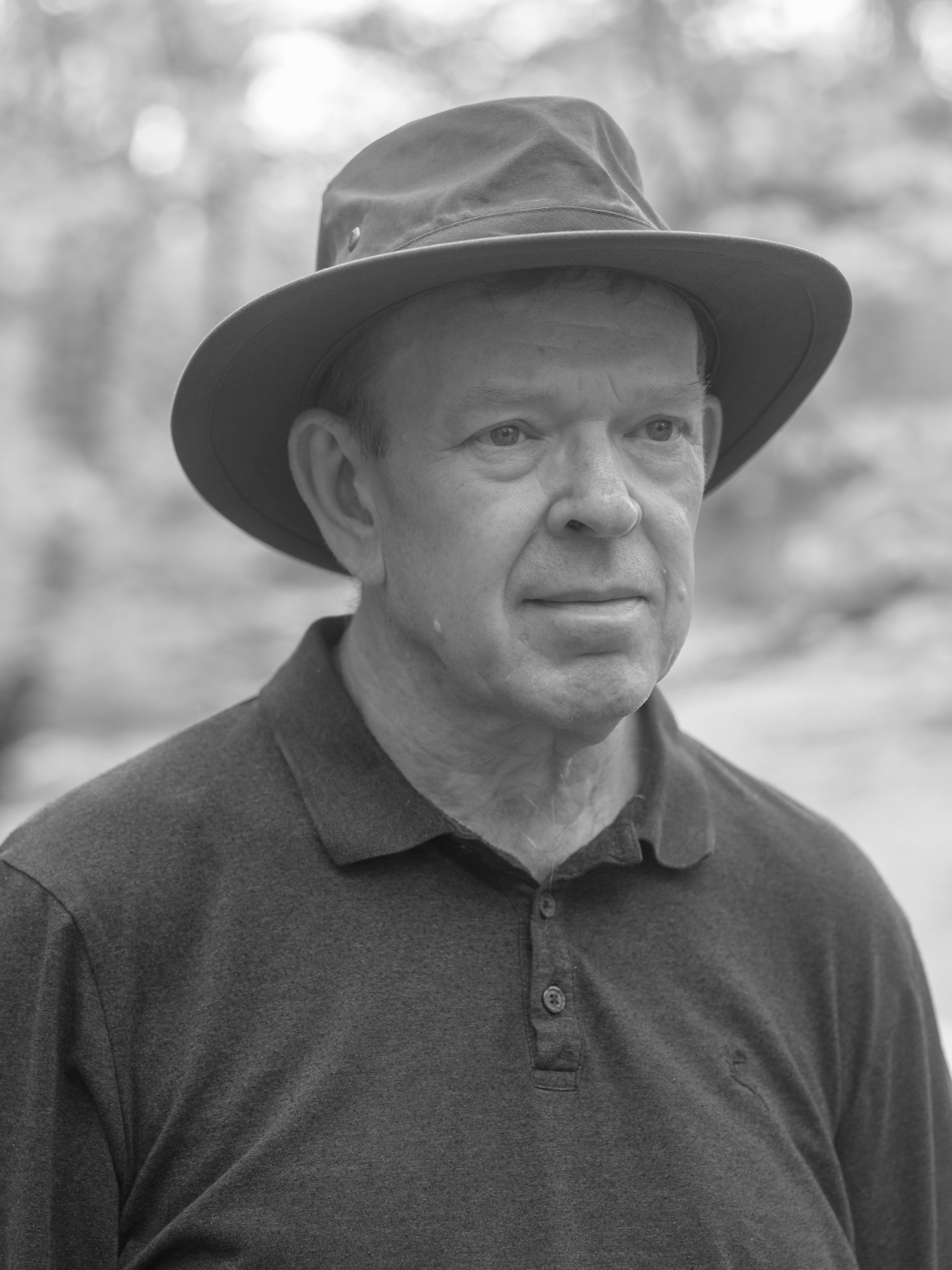 Black and white photo of a middle-aged man wearing a wide-brimmed hat and a dark polo shirt, outdoors with blurred trees in the background.