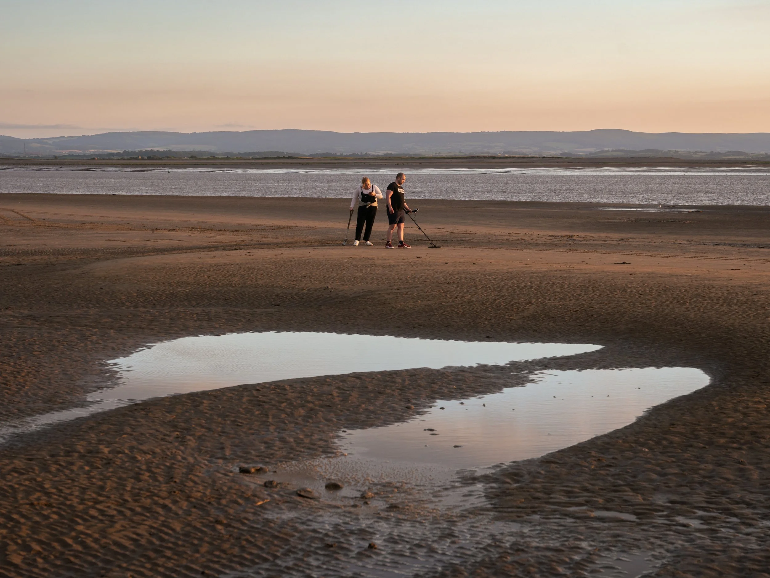 Two people are on a sandy beach with a large heart-shaped water pool in the foreground, looking at the ground in the distance during sunset, with water and hills in the background.