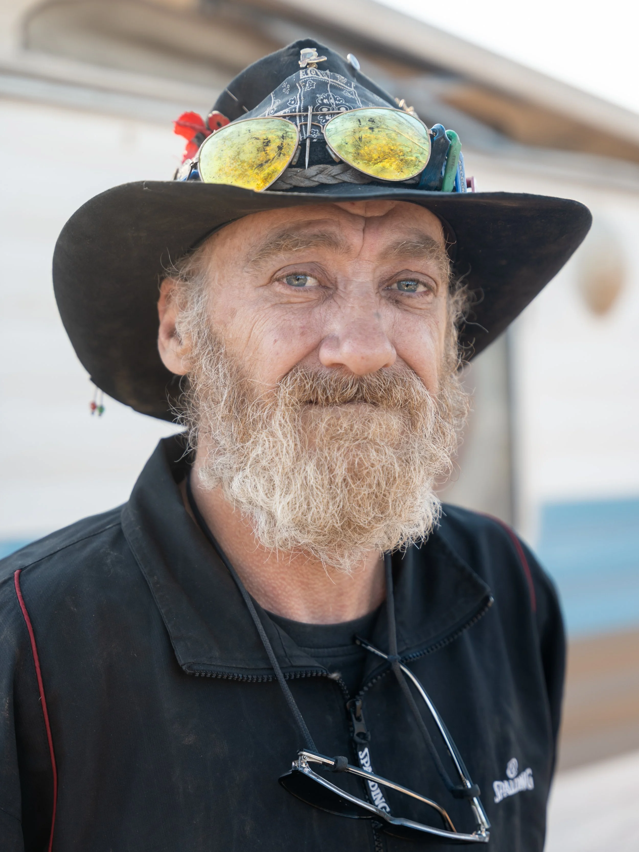 Close-up of an older man with a beard wearing a wide-brimmed hat adorned with reflective sunglasses and various small items, dressed in a black jacket.