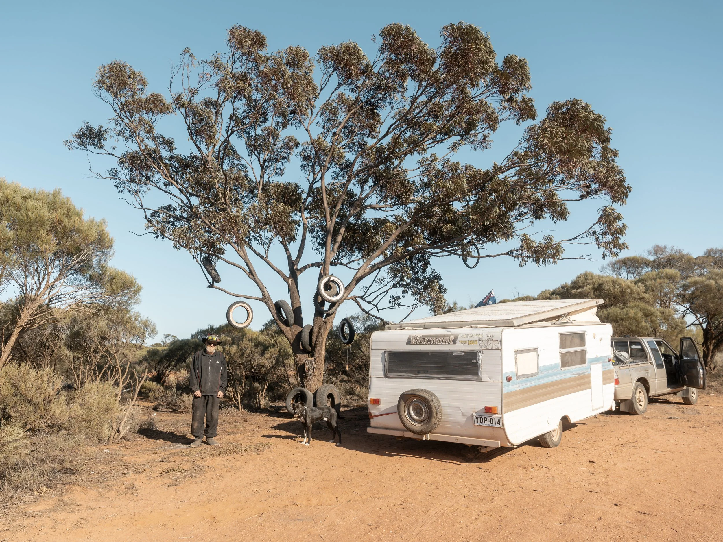 A man standing next to a black dog beside a tree decorated with tires, on a dirt campsite with a white camper trailer and a pickup truck in a dry, bushy area under a clear blue sky.