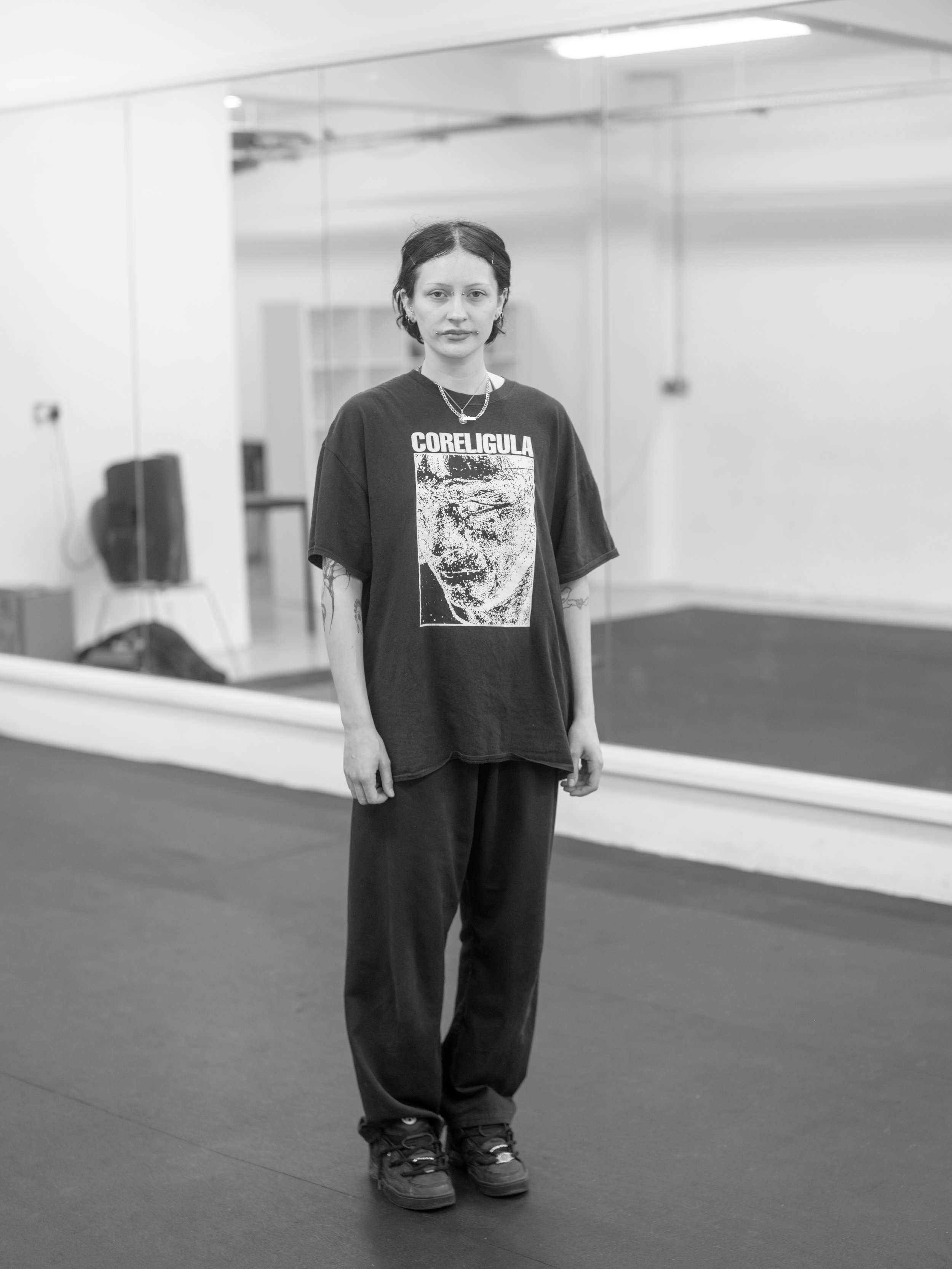 A young woman standing in a dance studio or gym, wearing a graphic T-shirt, loose pants, and sneakers, with a mirror and exercise equipment in the background.